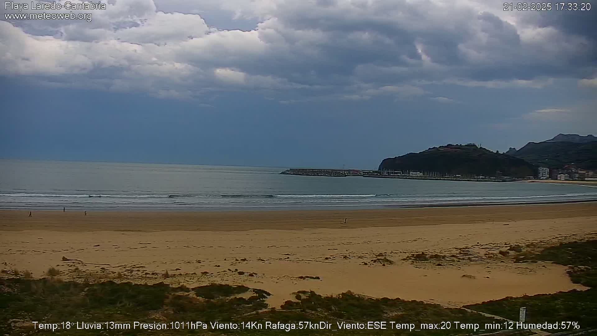 A mostly cloudy day shows a wide sandy beach with a calm ocean, a small headland and town in the distance.