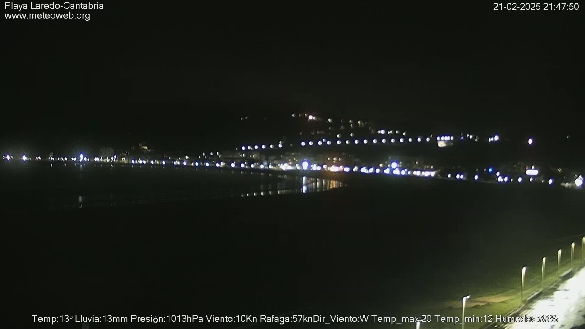 A nighttime long shot of a coastal town illuminated by lights, with rain.