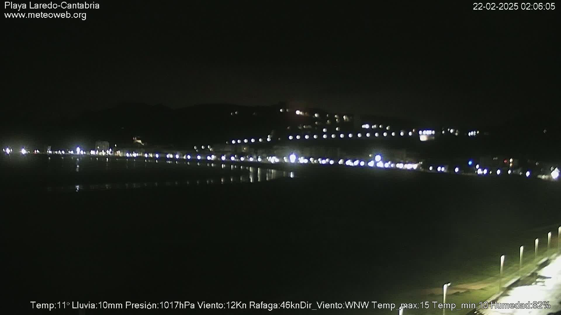 A nighttime view of a dark, rain-soaked beach with a line of lights from buildings visible in the distance.