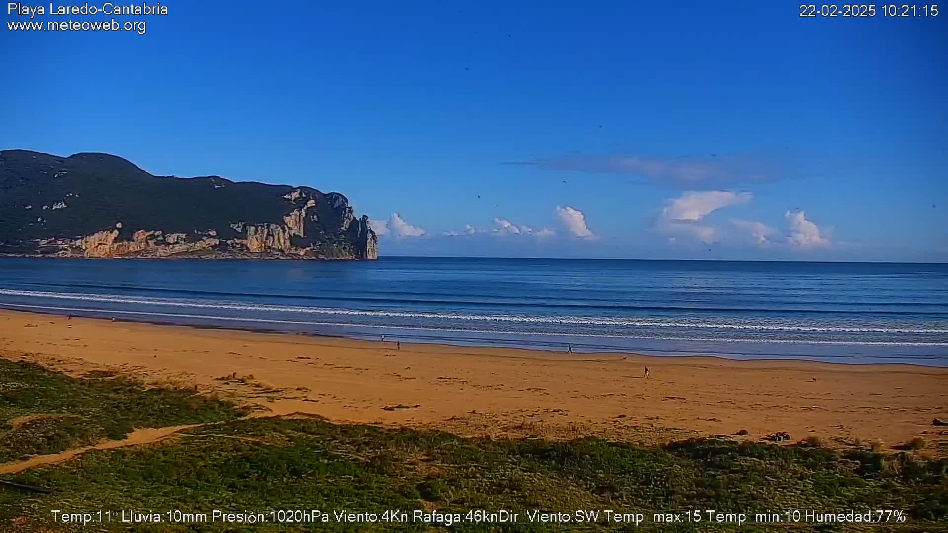A sandy beach stretches toward a calm ocean under a mostly clear blue sky with scattered clouds, a dark green and rocky headland visible in the distance.