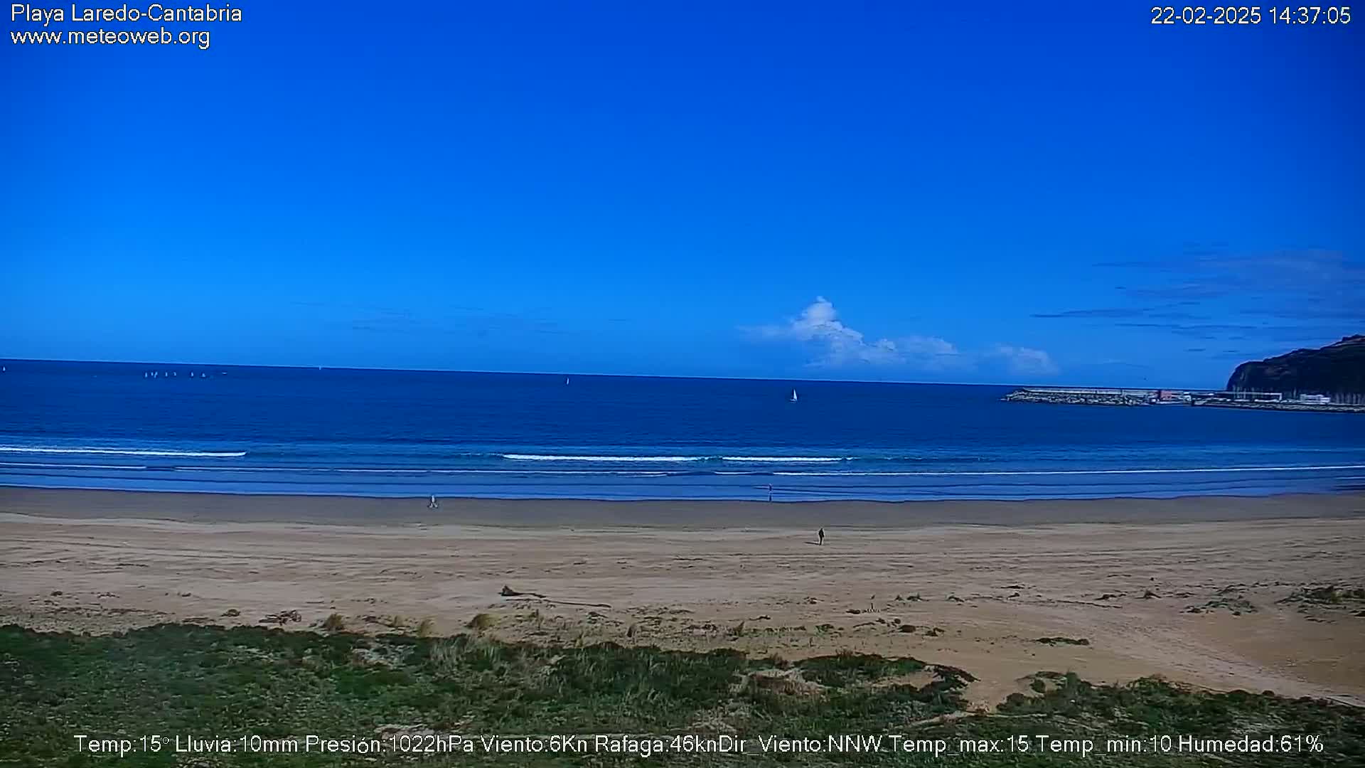 A sandy beach stretches to a calm, blue ocean under a mostly clear blue sky with a few small clouds, and a distant headland is visible to the right.
