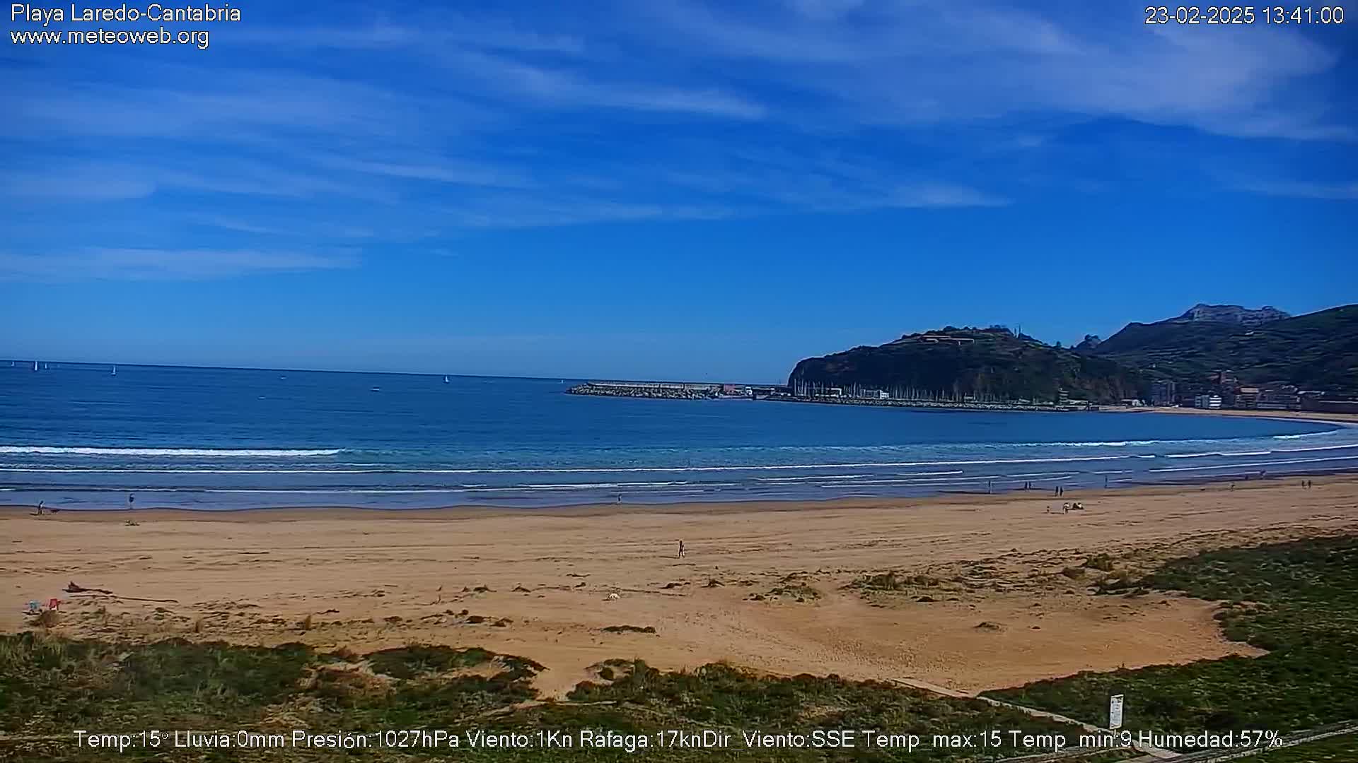 A sandy beach with a calm ocean, small waves, and a few people, is situated below a hill with buildings under a mostly clear blue sky.