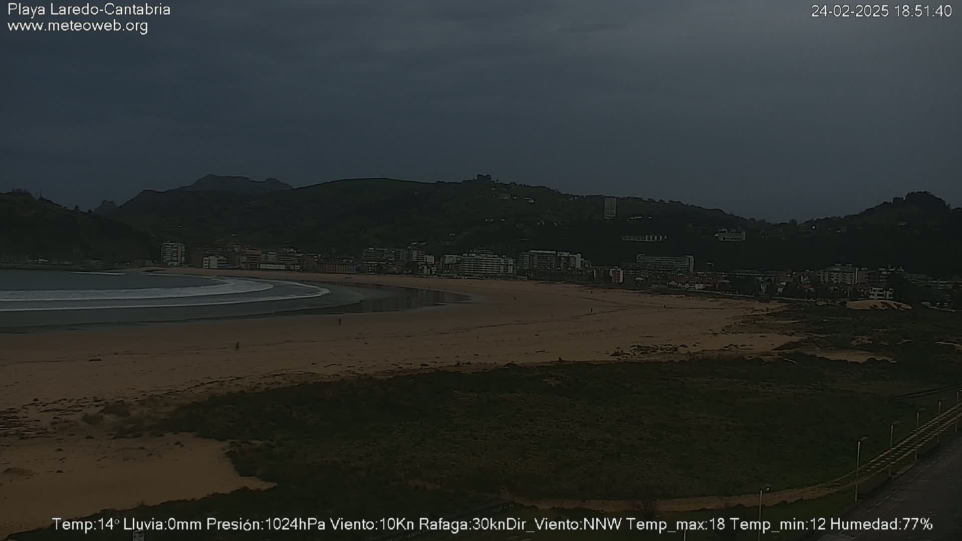 A sandy beach curves along a bay under a dark, overcast sky, with a town and hills visible in the background.