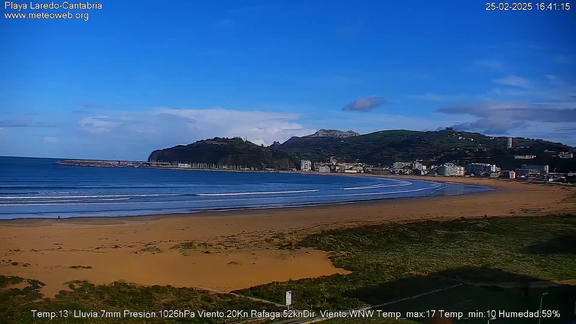 A sandy beach with gentle waves laps the shore under a mostly clear blue sky, with a town and hills visible in the background.