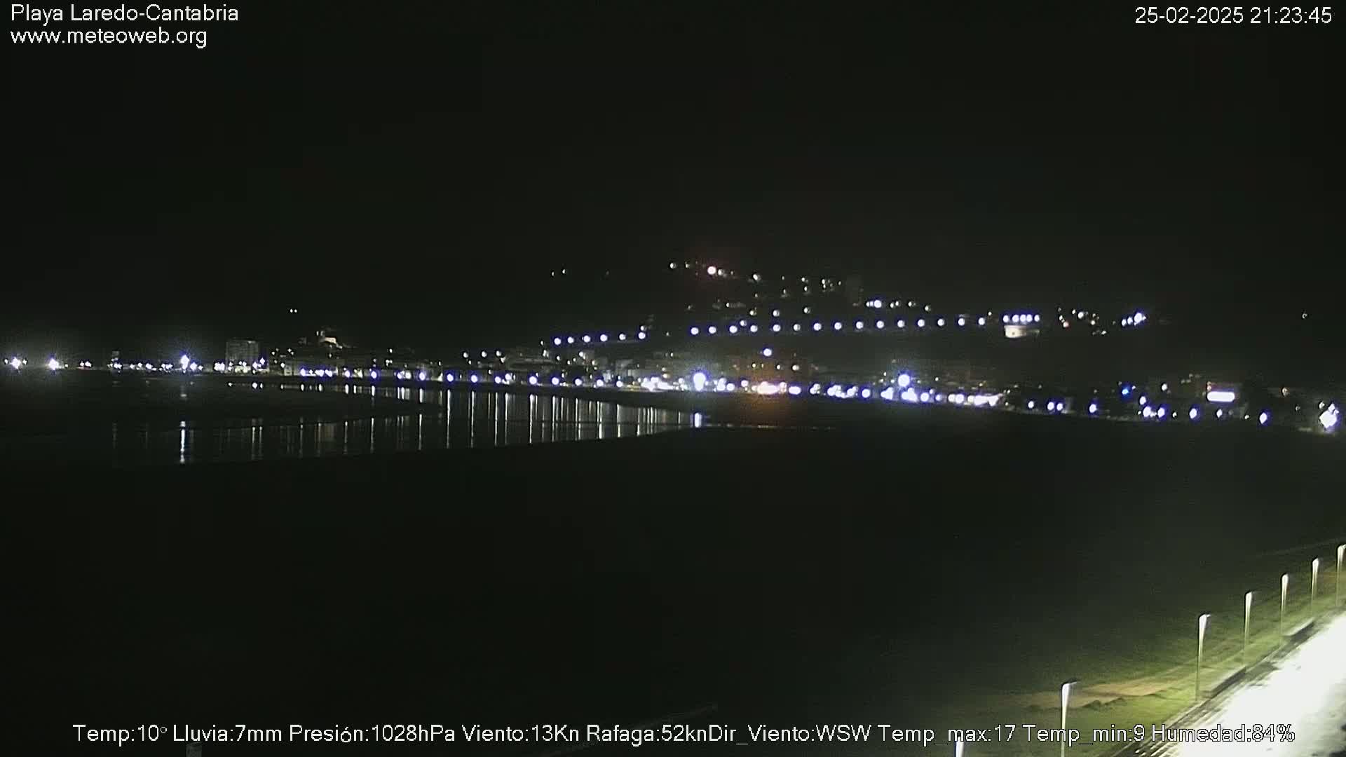 A nighttime view of a coastal town's lights reflected in dark water, under a dark sky with rain.