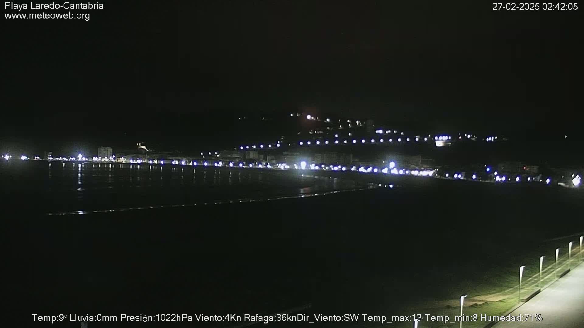 A nighttime view of a dimly lit beach town across a dark body of water, under clear skies.