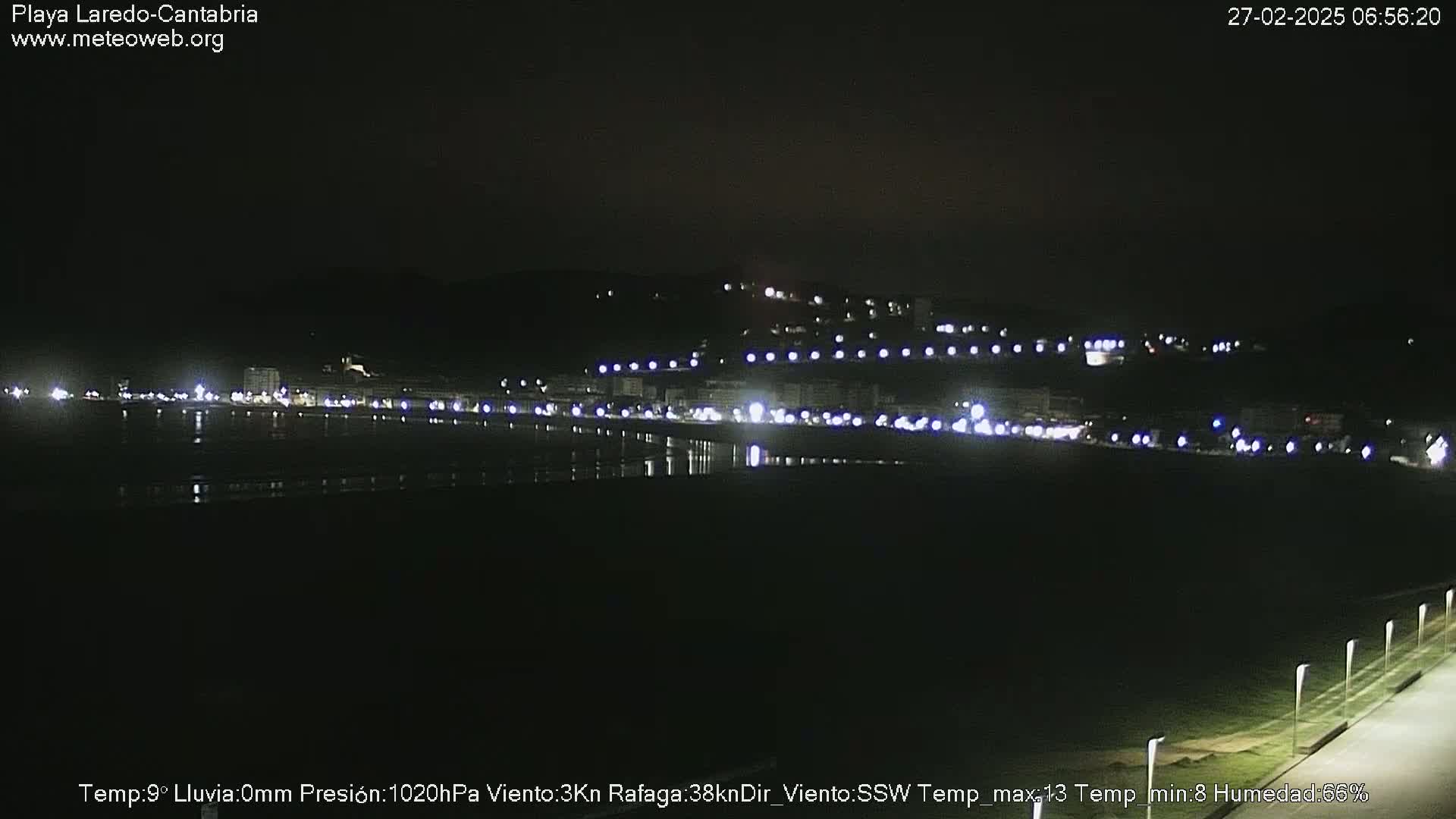 A nighttime long shot of a coastal town with lights reflecting on the water under a clear, dark sky.