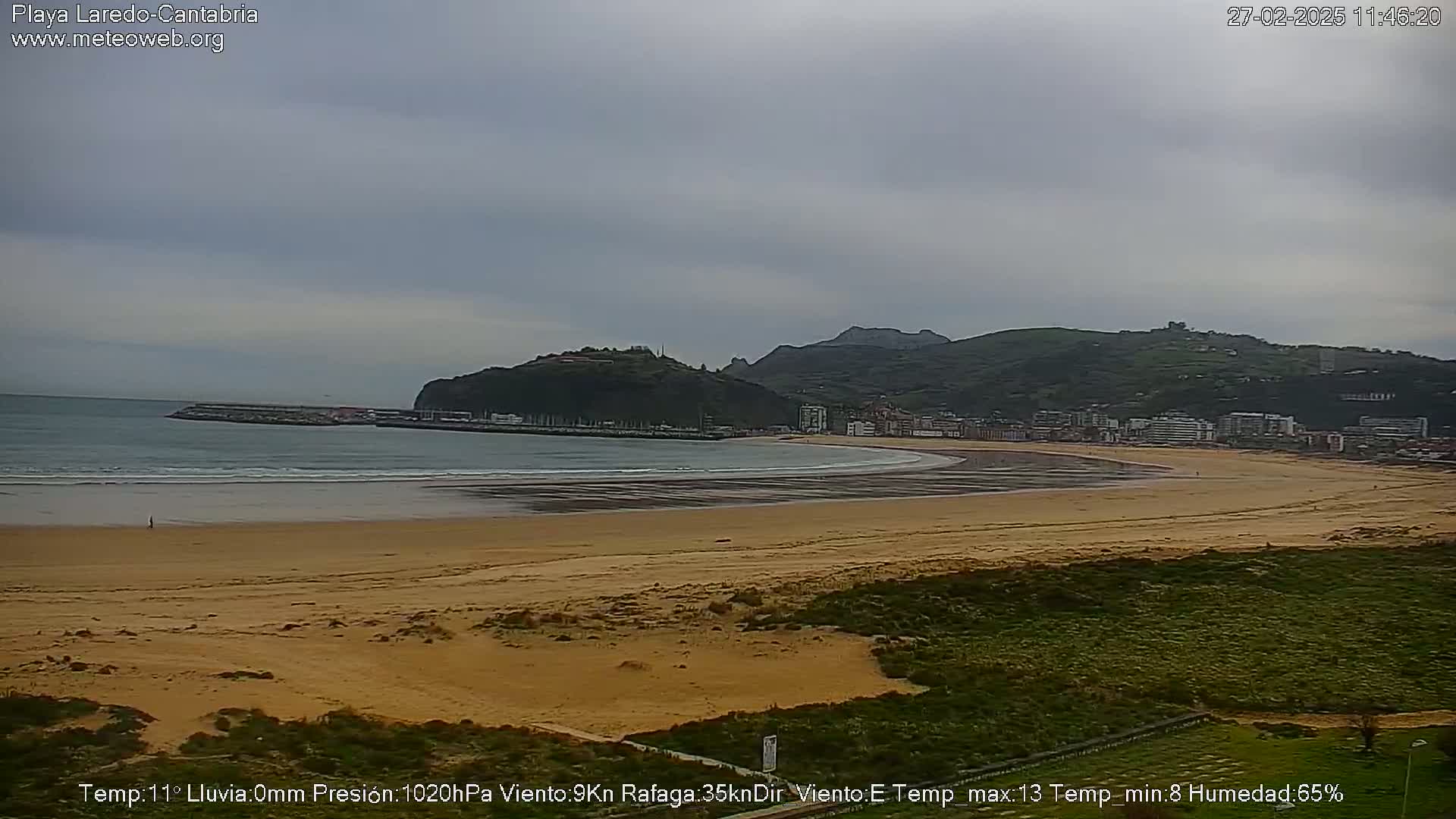 A long sandy beach curves gently around a bay, backed by a town nestled against a hillside under a mostly cloudy sky.