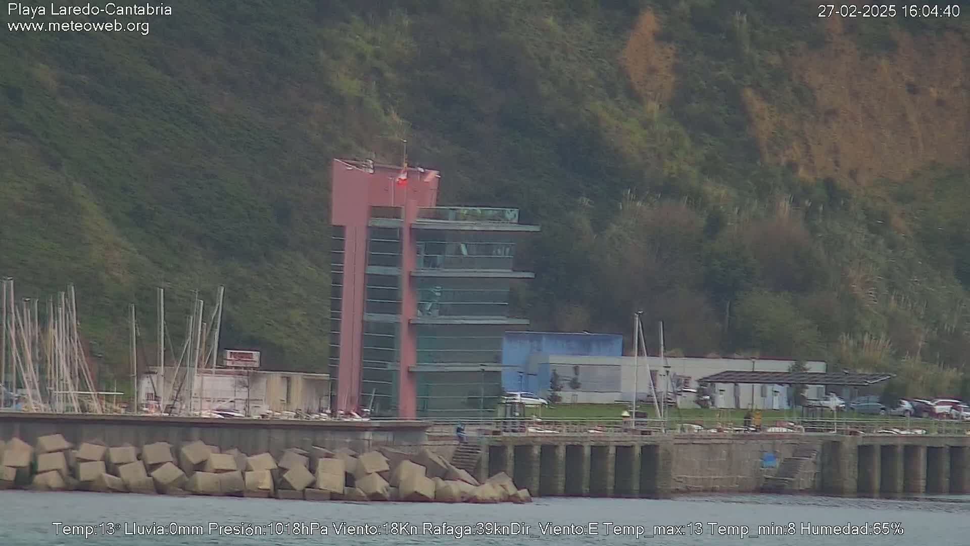A pink multi-story building stands near a harbor with a breakwater, on a cloudy day.