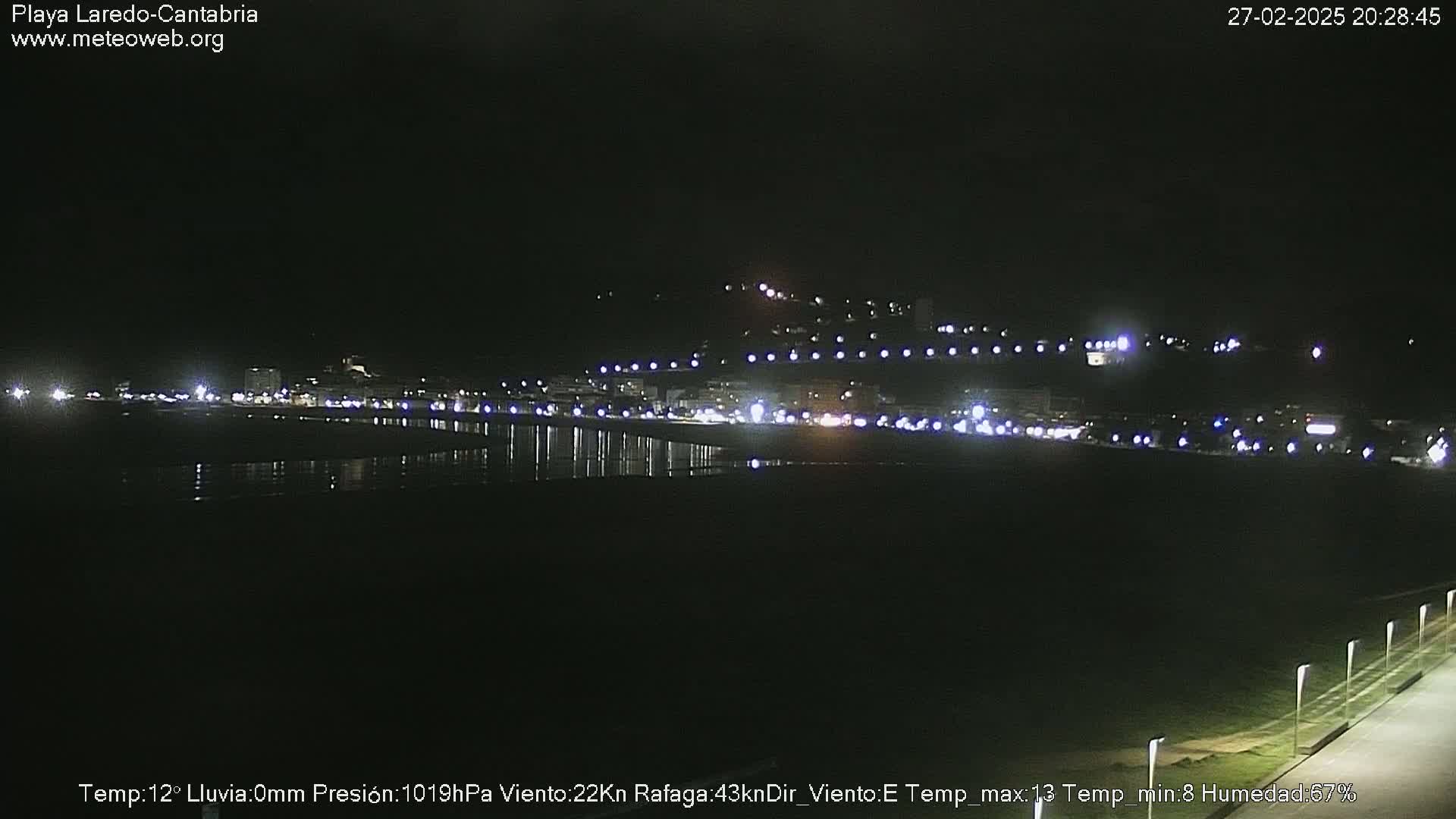A nighttime view of a coastal town with lights reflecting on the water, under a clear, dark sky.