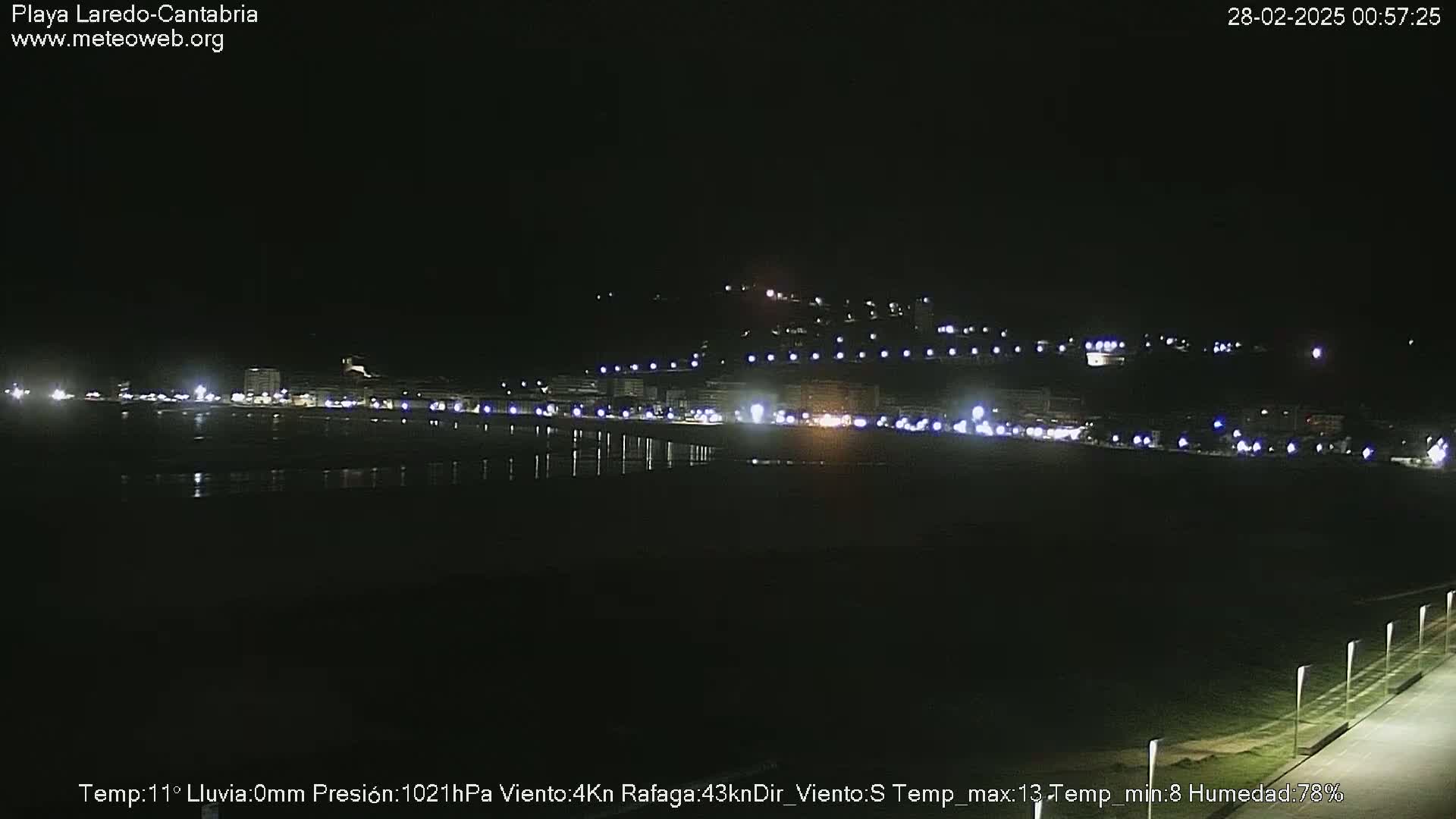 A nighttime view of a dark, calm beach with a town's lights illuminating the shoreline in the distance.