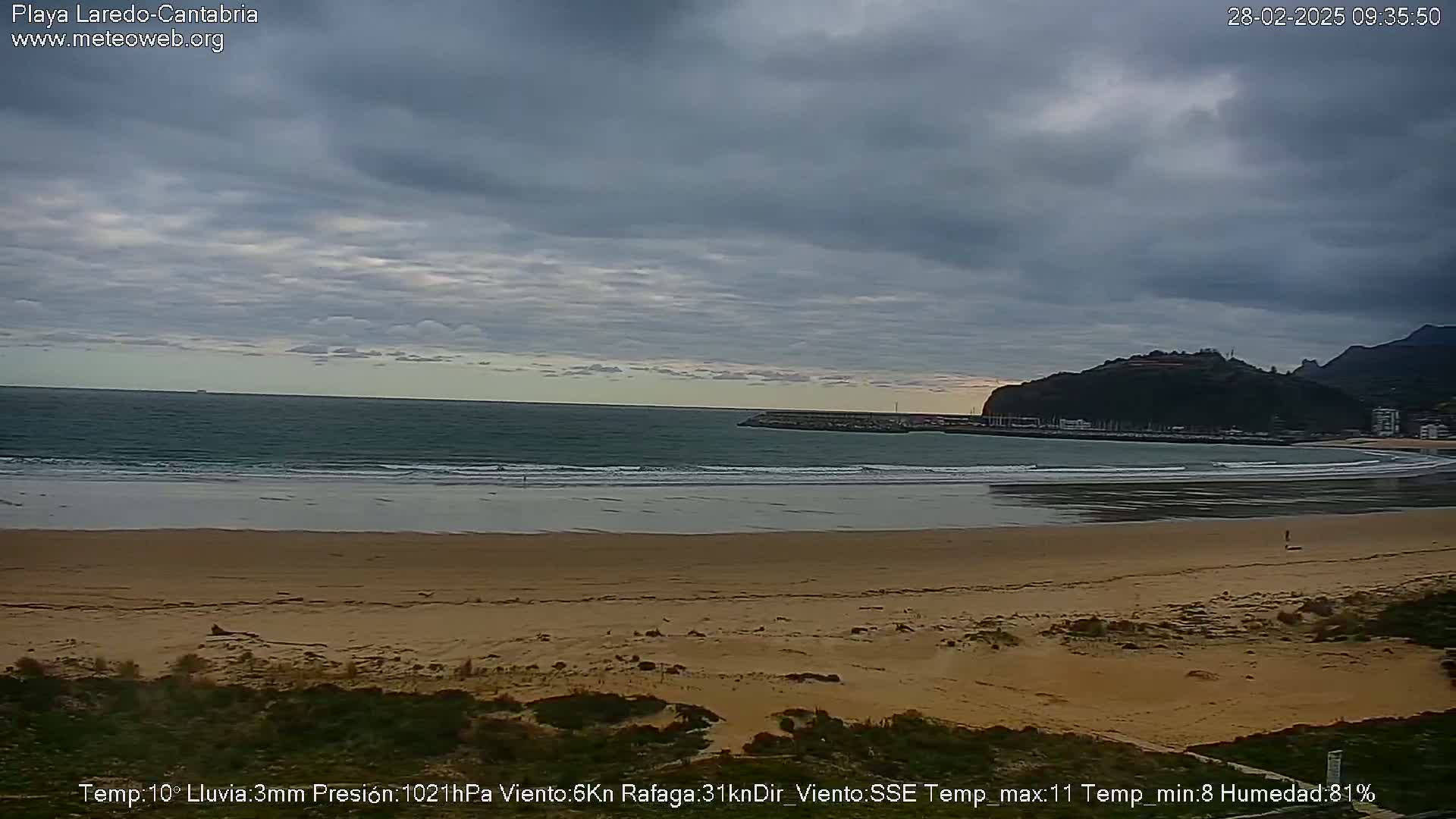 A sandy beach with gentle waves meets a calm ocean under an overcast sky.