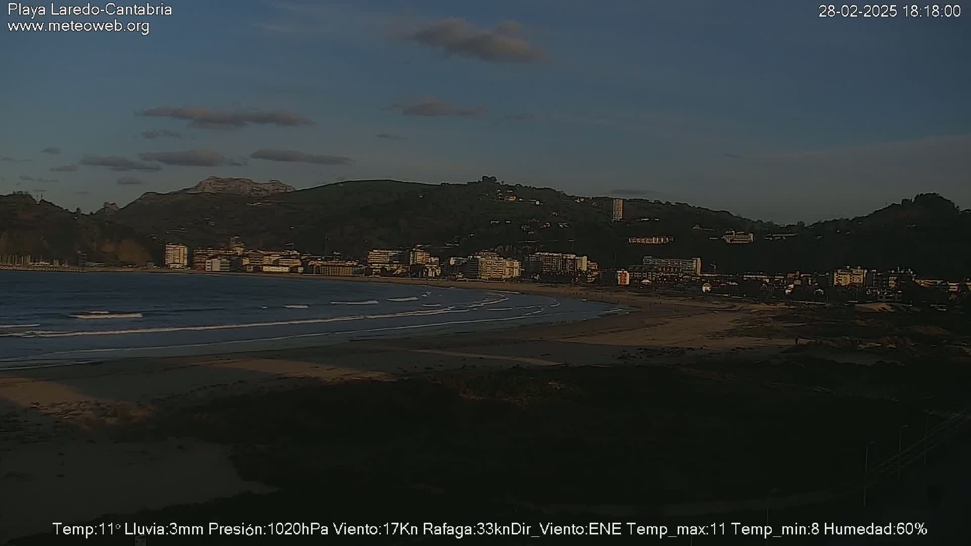 A beach at dusk with small waves, backed by a town nestled against hills under a mostly clear sky.