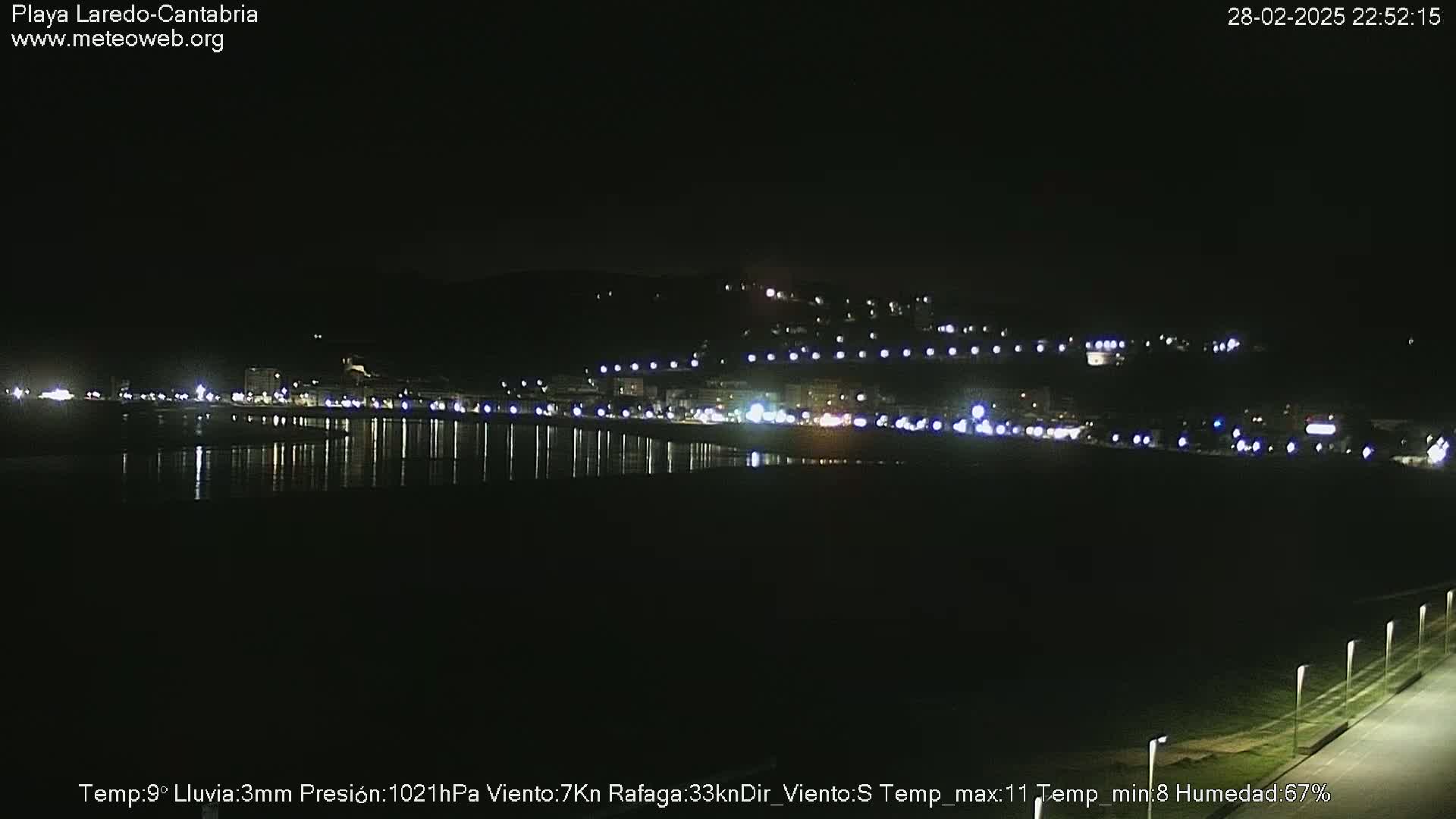 A nighttime coastal scene shows a dark beach with a town's lights reflecting on calm water under a clear, dark sky.