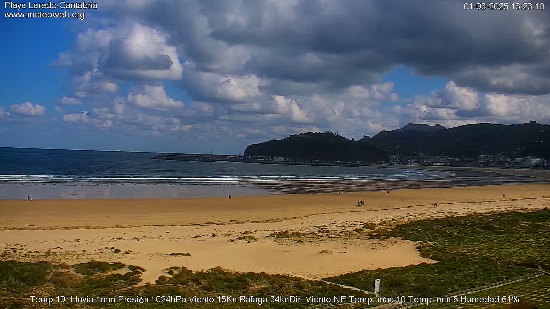 A mostly cloudy day at a sandy beach with small waves, a calm ocean, and a hill in the background.