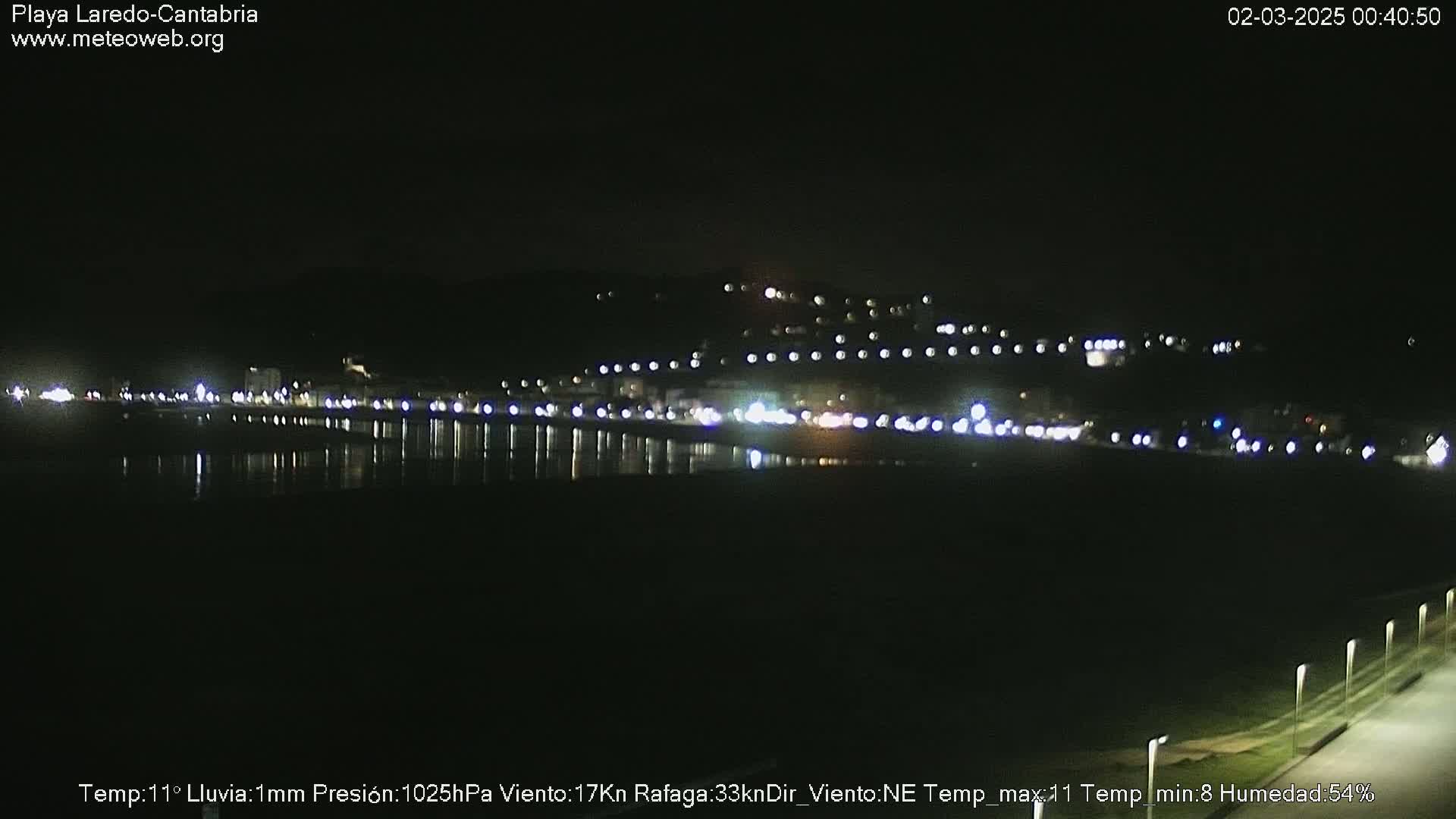 A nighttime view of a dark, calm body of water reflecting the lights of a town on a hill in the distance, under a clear sky.