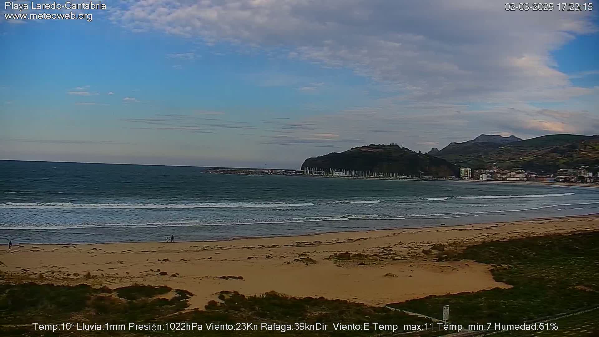 A sandy beach with gentle waves laps the shore under a partly cloudy sky; a town and hills are visible in the background.