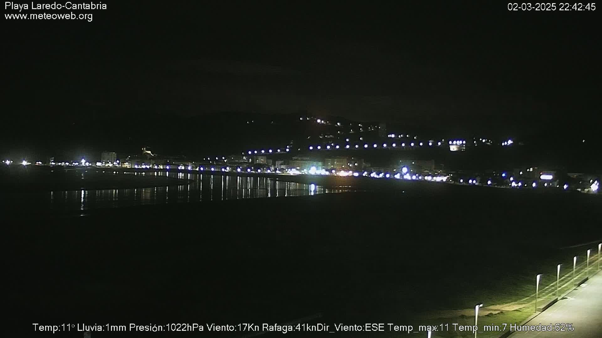 A nighttime coastal scene shows a town's lights reflecting on calm water under a dark sky with light rain.
