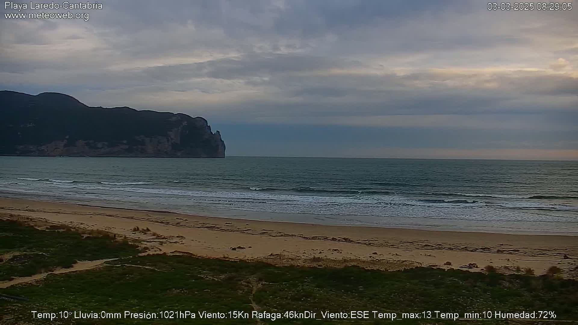 A sandy beach with small waves laps the shore under an overcast sky, with a dark mountain visible in the distance.