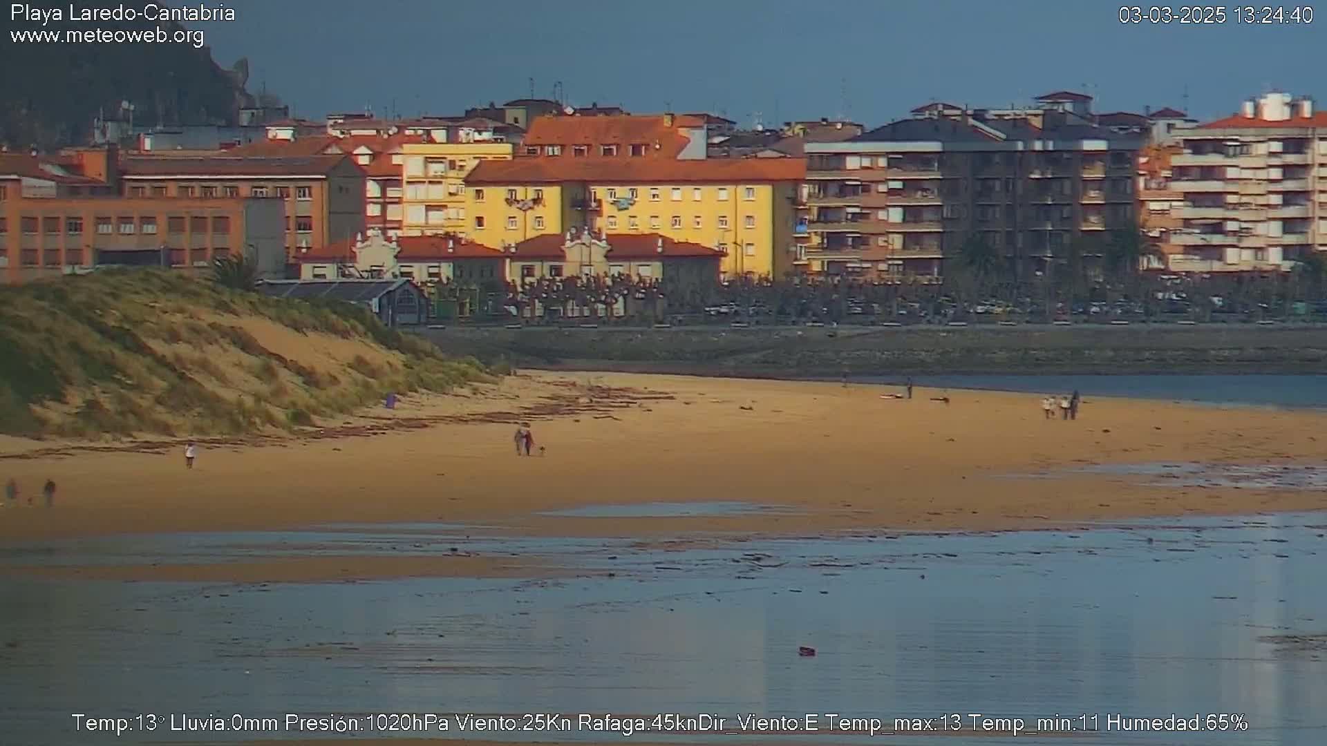 A sandy beach with a few people walking on it stretches out to calm water in front of a row of multi-story buildings under a cloudy sky.