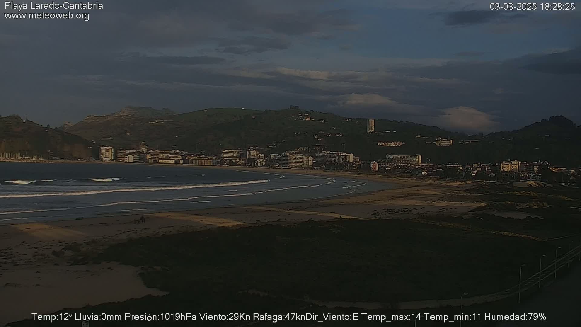 A beach with small waves under a cloudy sky overlooks a town nestled against a hill.