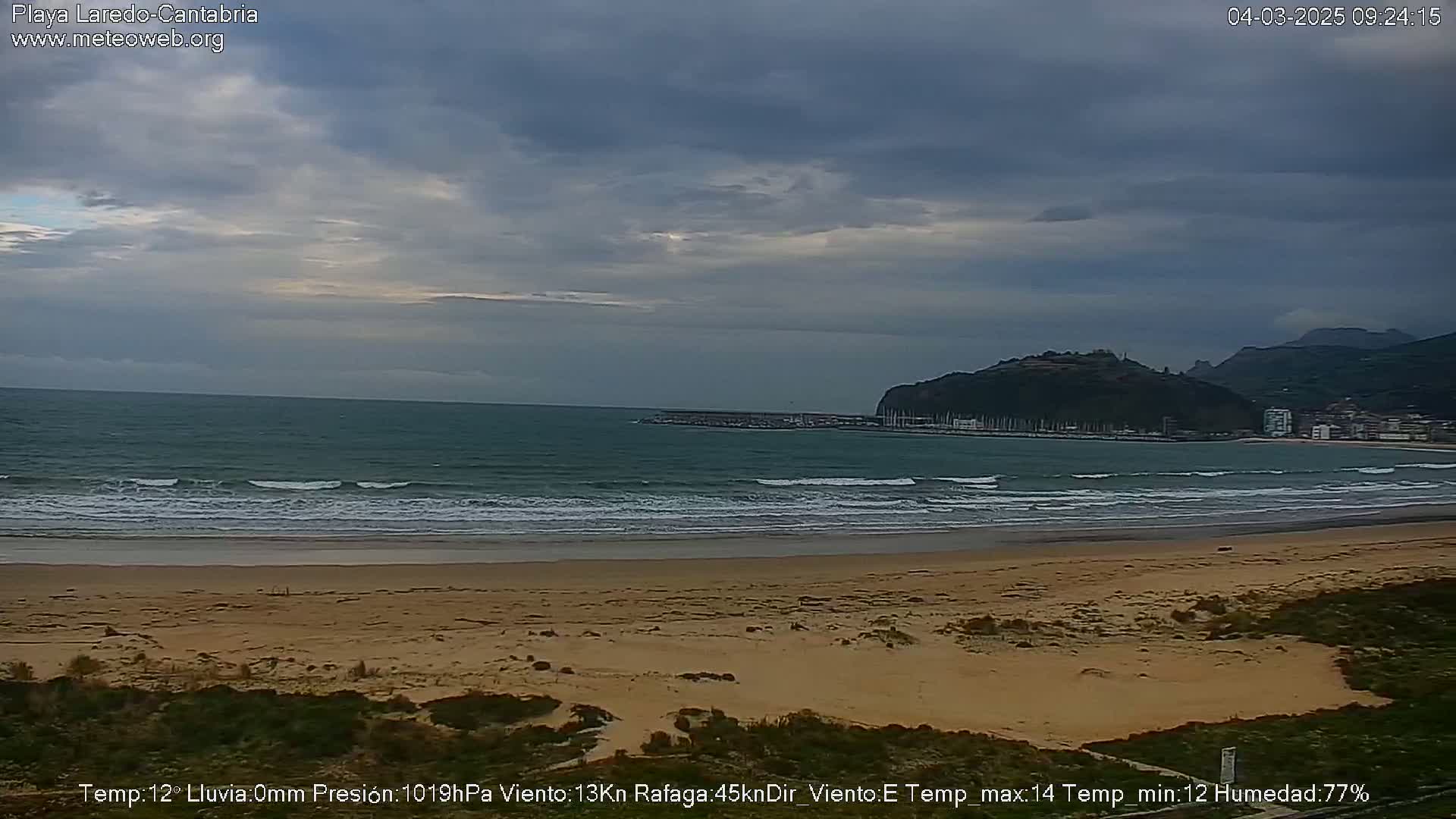 A sandy beach with small waves under an overcast sky overlooks a calm bay and distant hills.