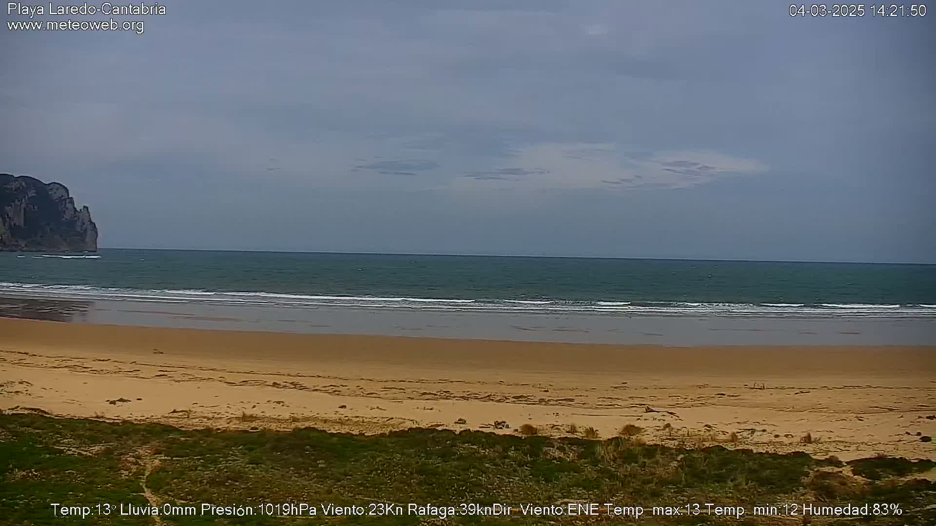 A sandy beach stretches to a calm, teal ocean under a mostly cloudy sky, with a rocky cliff visible in the distance.