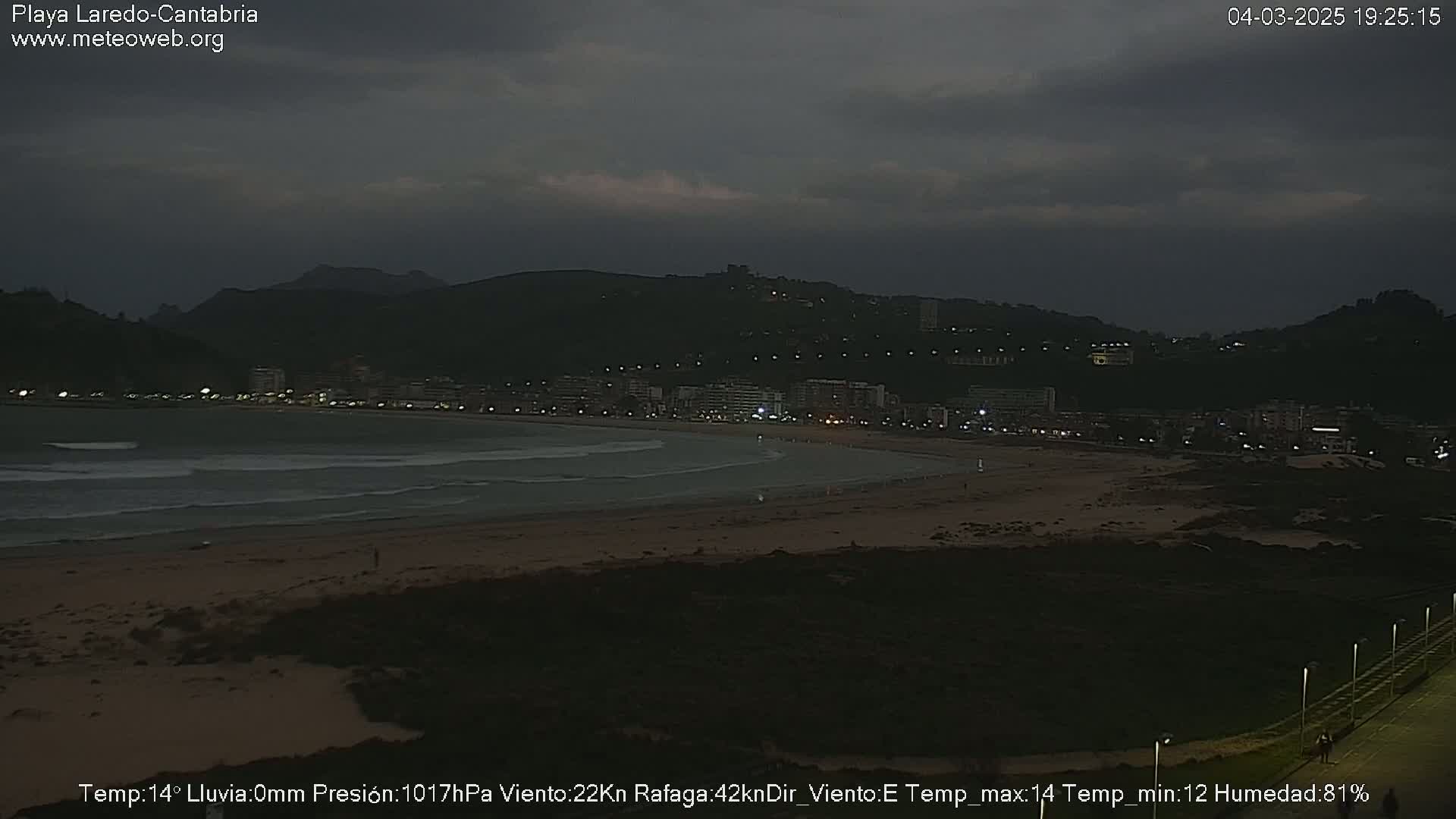 A dark, overcast beach scene at night shows a town nestled against a mountain, with waves lapping the shore.