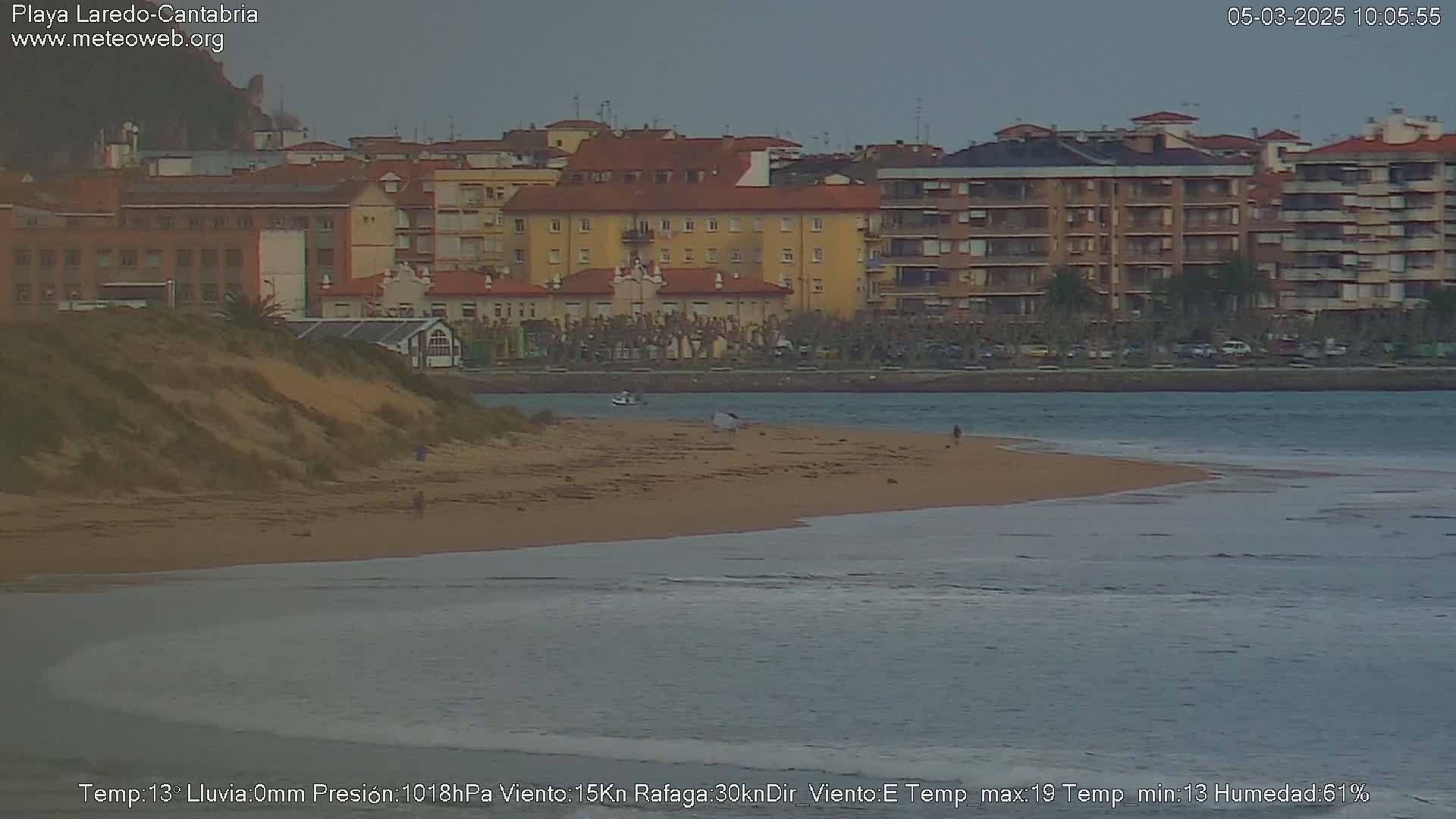 A sandy beach with a few people on it stretches along a calm body of water in front of a row of buildings under an overcast sky.