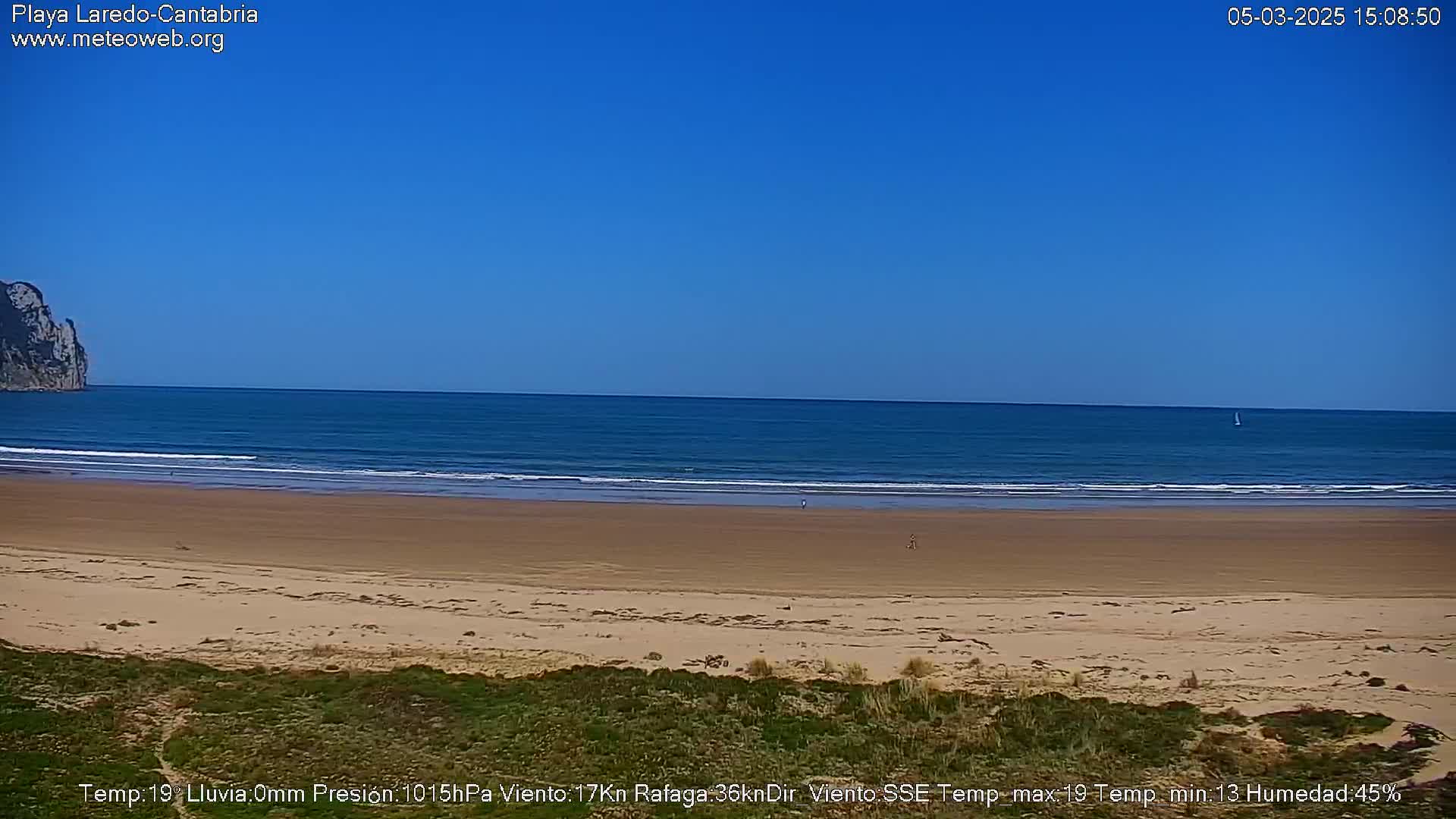 A sandy beach stretches to a calm, blue ocean under a clear blue sky; a lone sailboat is visible on the horizon.