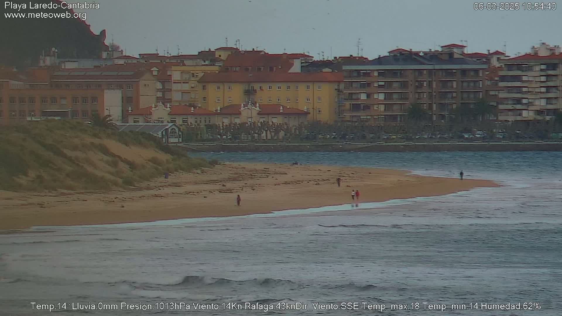 A sandy beach with a few people walking on it stretches out before a row of buildings under an overcast sky.
