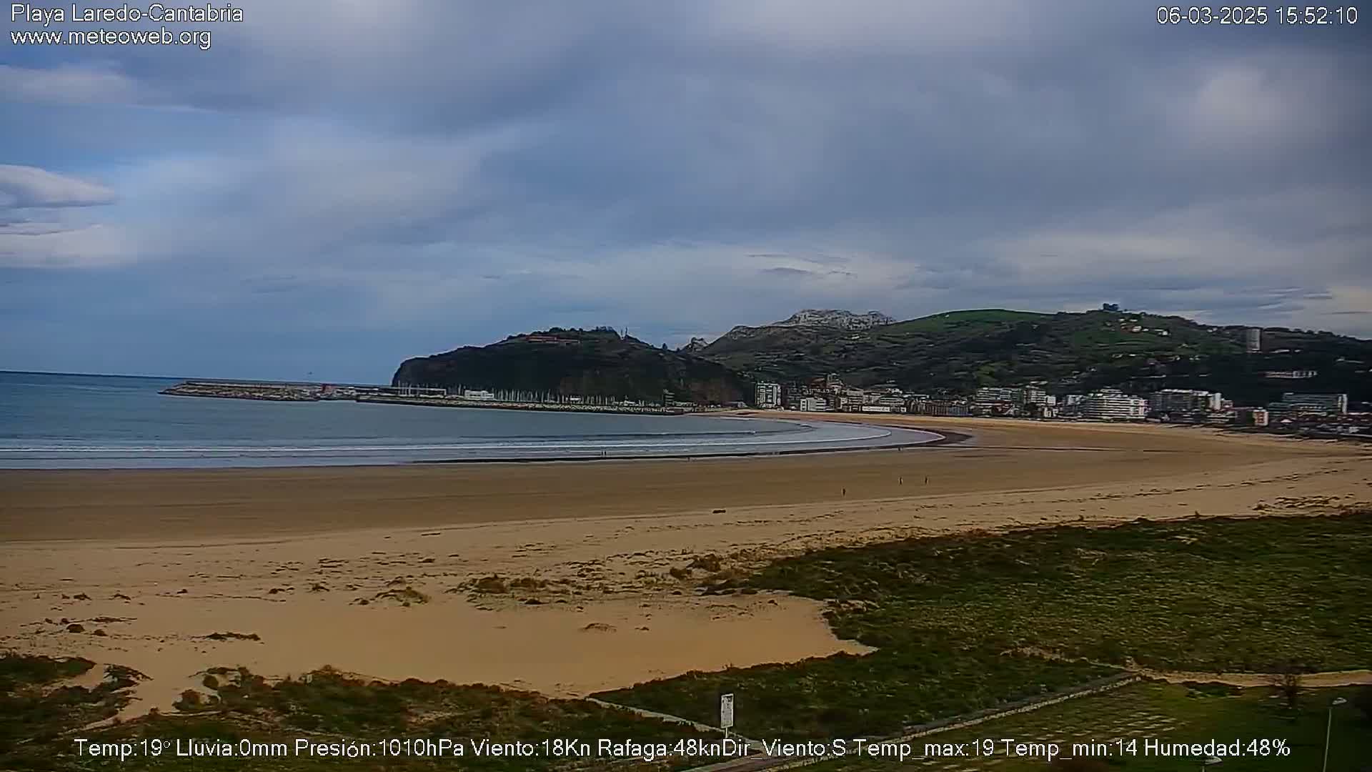 A wide sandy beach curves around a calm bay, with a town nestled against hills under a mostly cloudy sky.