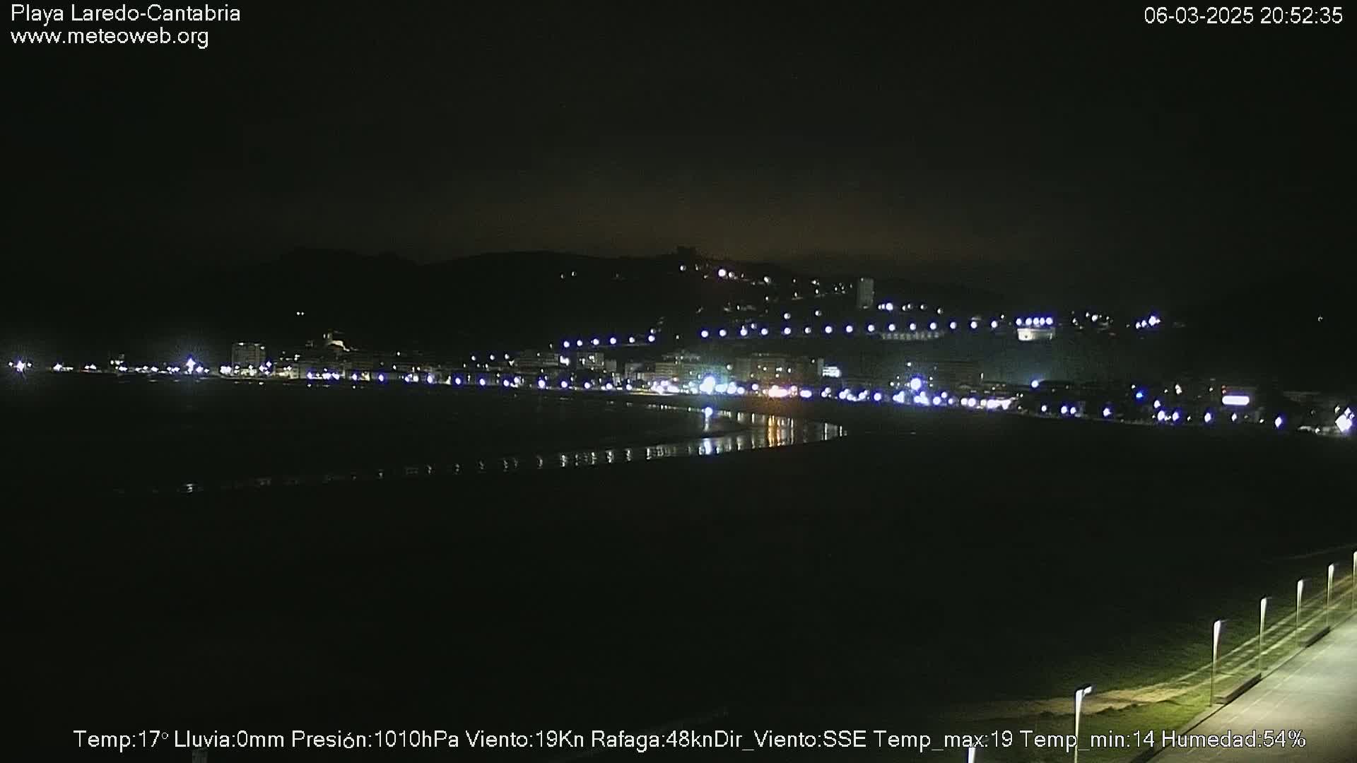 A nighttime view of a coastal town with its lights reflecting on a dark body of water under a clear, dark sky.