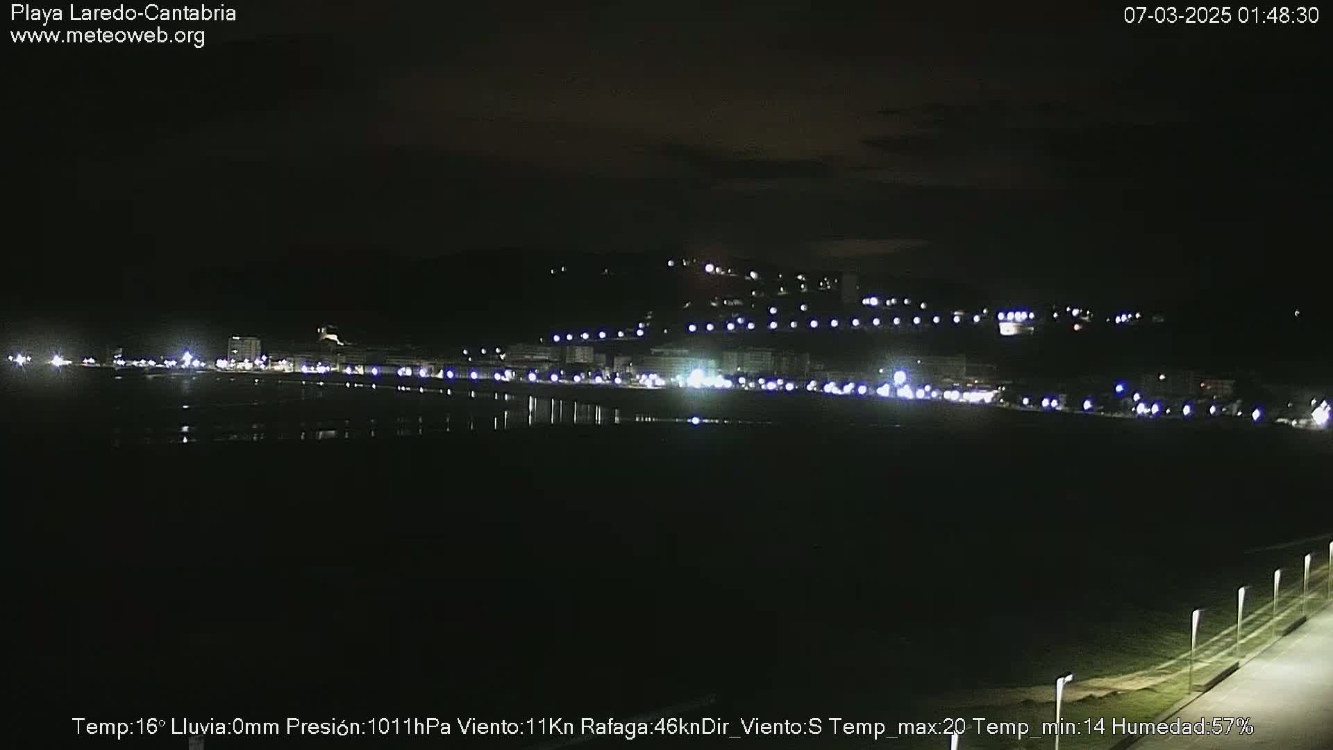 A nighttime view of a coastal town illuminated by lights, under a dark, clear sky.