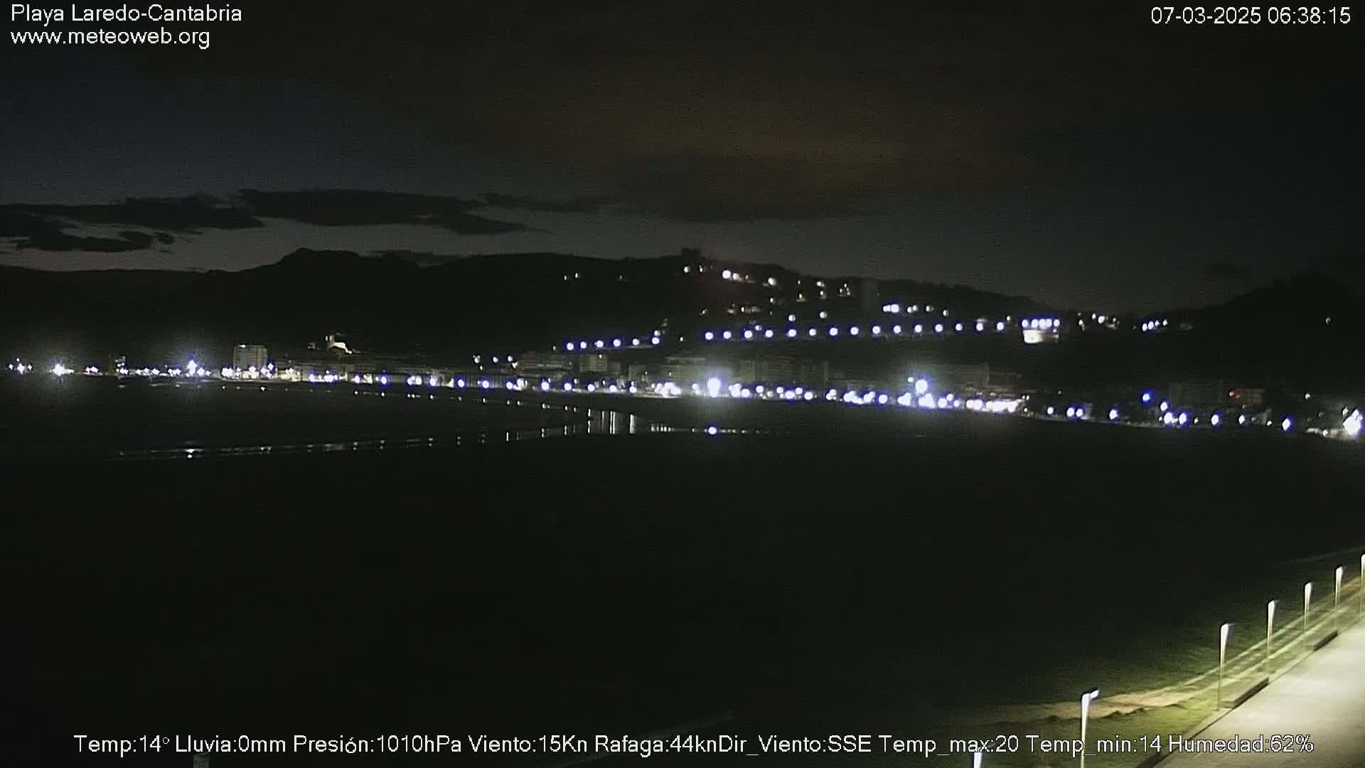 A nighttime view of a dark beach and dimly lit buildings along a hillside under a mostly clear sky.