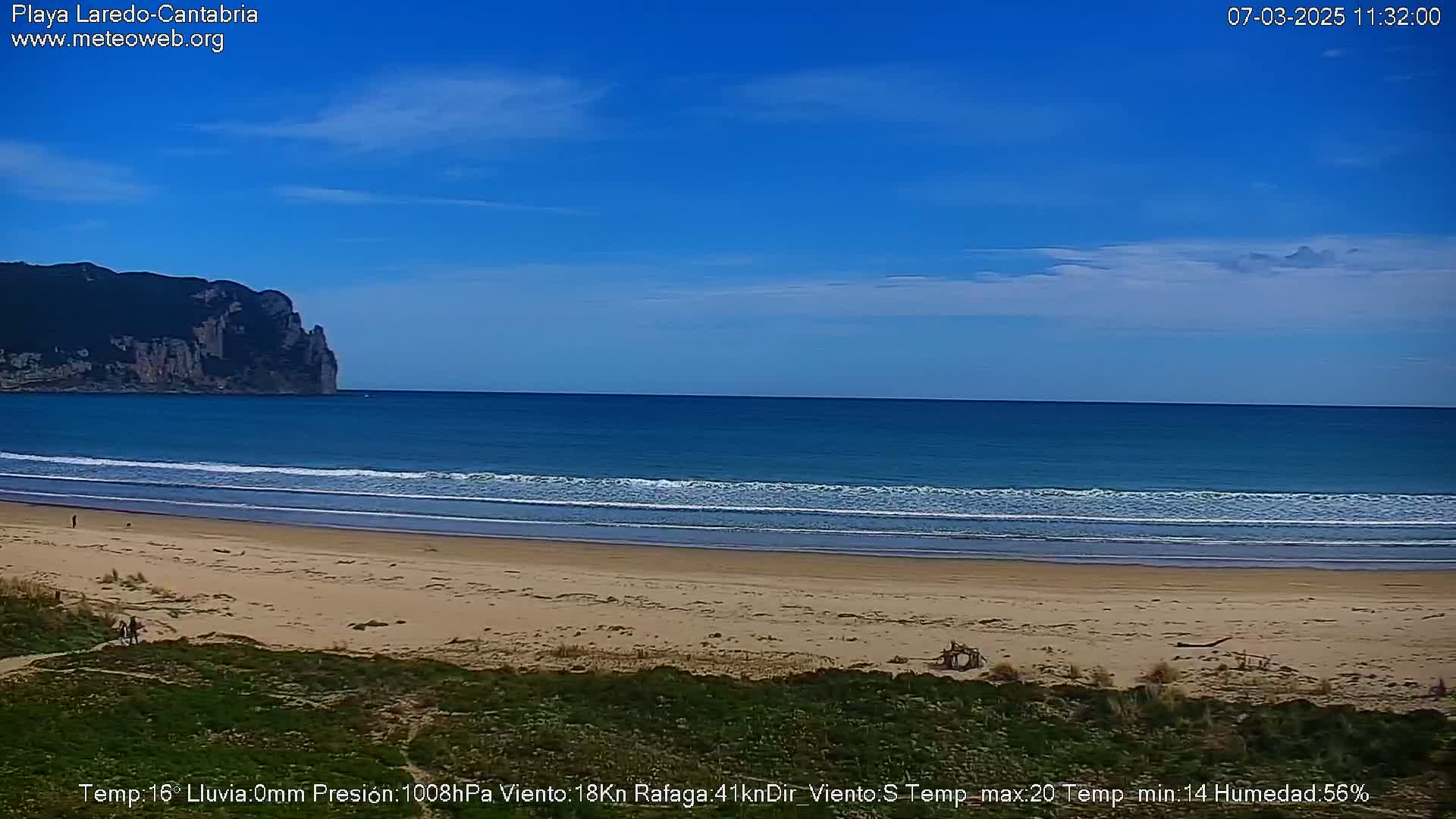 A sandy beach stretches beneath a clear blue sky, with gentle waves lapping the shore and a rocky cliff visible in the distance.