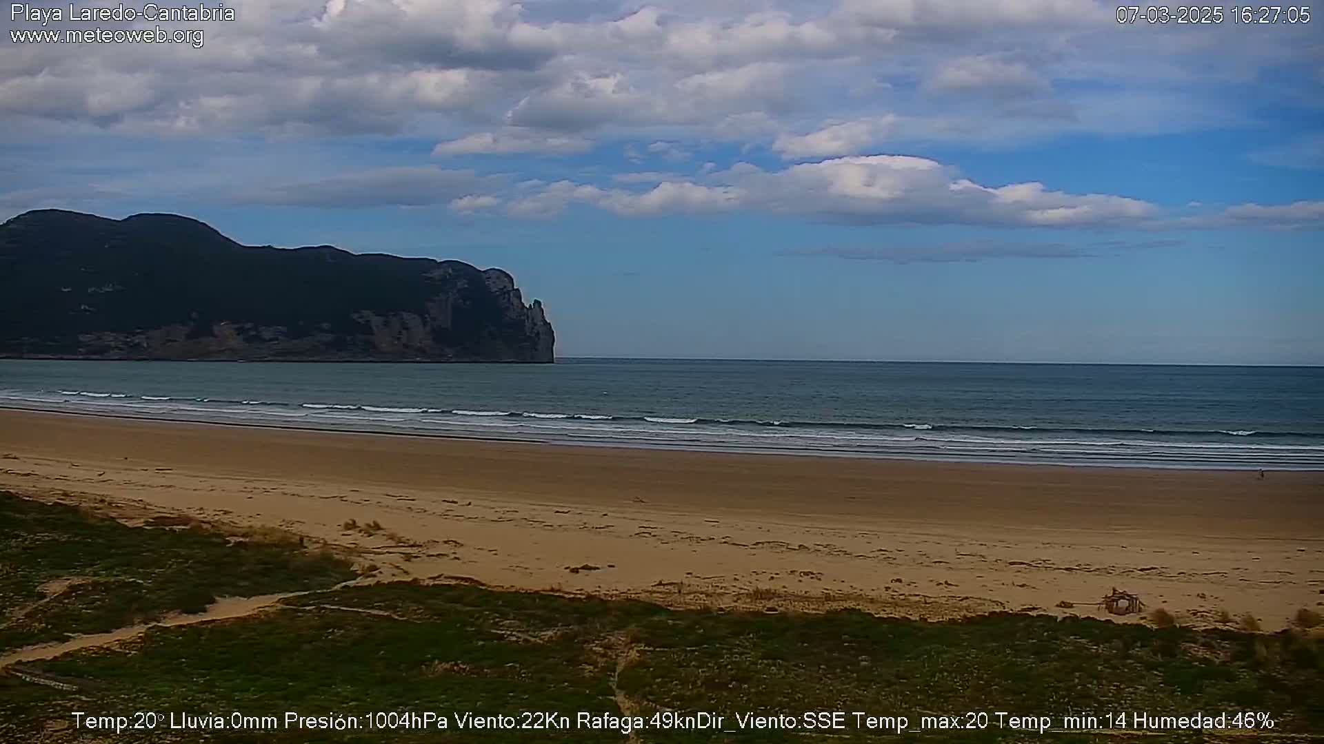 A sandy beach with small waves laps against the shore, under a partly cloudy sky, with a large, dark mountain visible in the distance.
