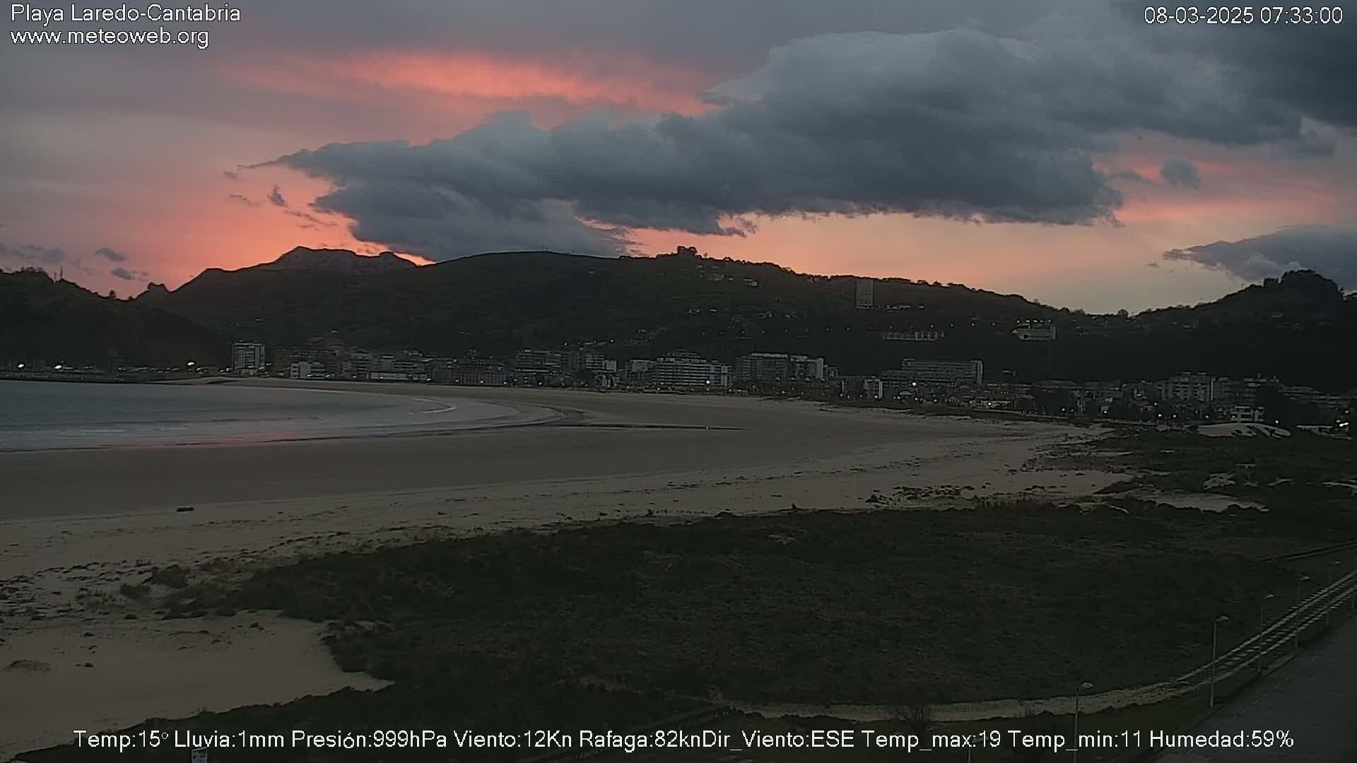 A sandy beach stretches before a town nestled at the base of a mountain range under a partly cloudy sky with pink hues near the horizon.