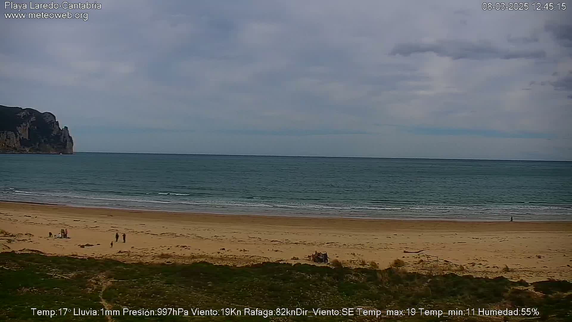 A sandy beach with a few people on it stretches to a calm sea under a cloudy sky, with a cliff visible in the distance.