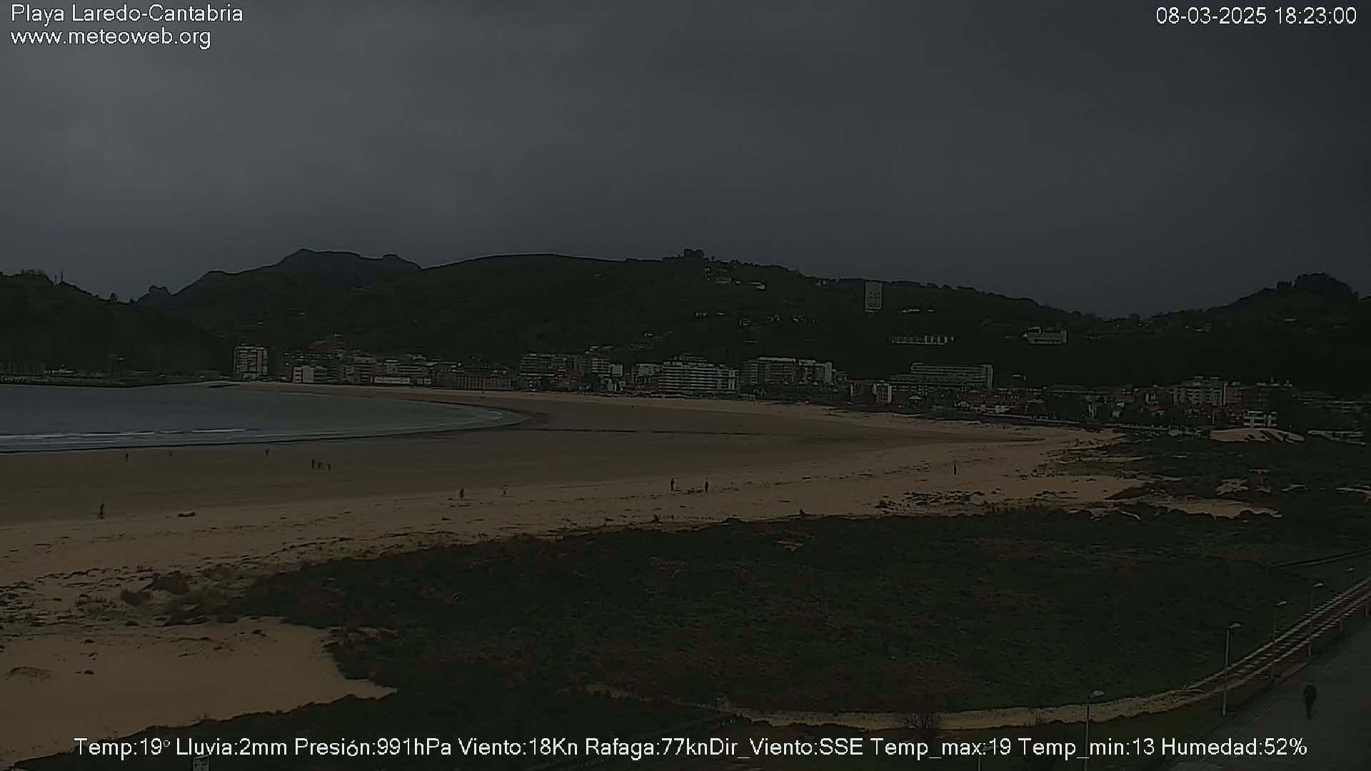 A beach with a town and hills in the background under an overcast sky.