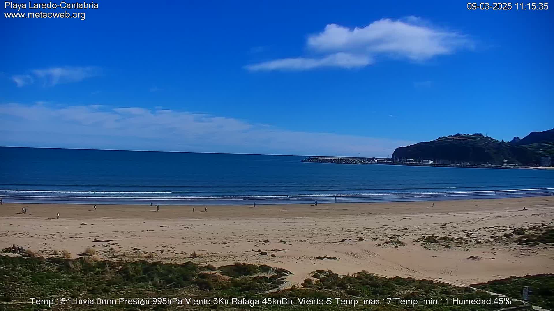 A mostly sunny day at a sandy beach with a calm ocean and a few people walking on the sand, with a distant hill visible in the background.