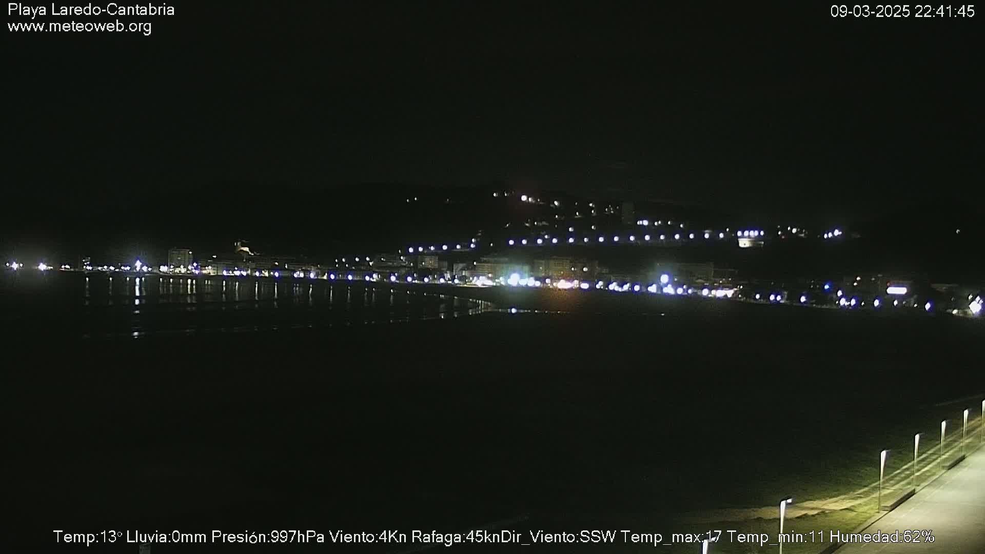 A nighttime view of a dimly lit coastal town with a dark beach and calm water reflecting the lights.