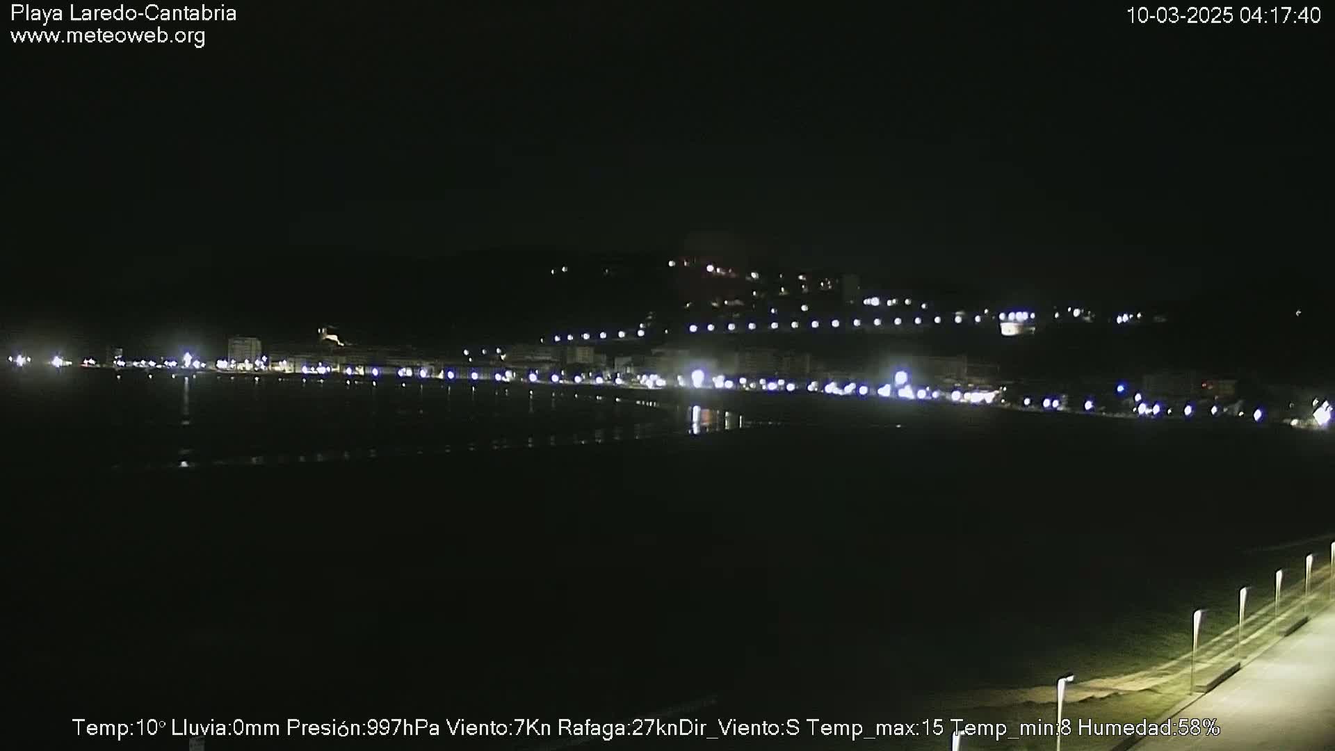 A nighttime view of a dark, calm body of water reflecting the lights of a town on a hill in the distance.