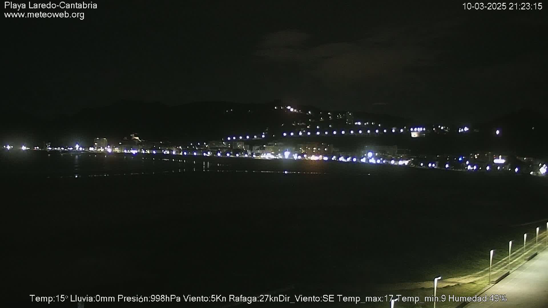 A nighttime view of a dimly lit beach town, across a dark body of water, under a clear, dark sky.