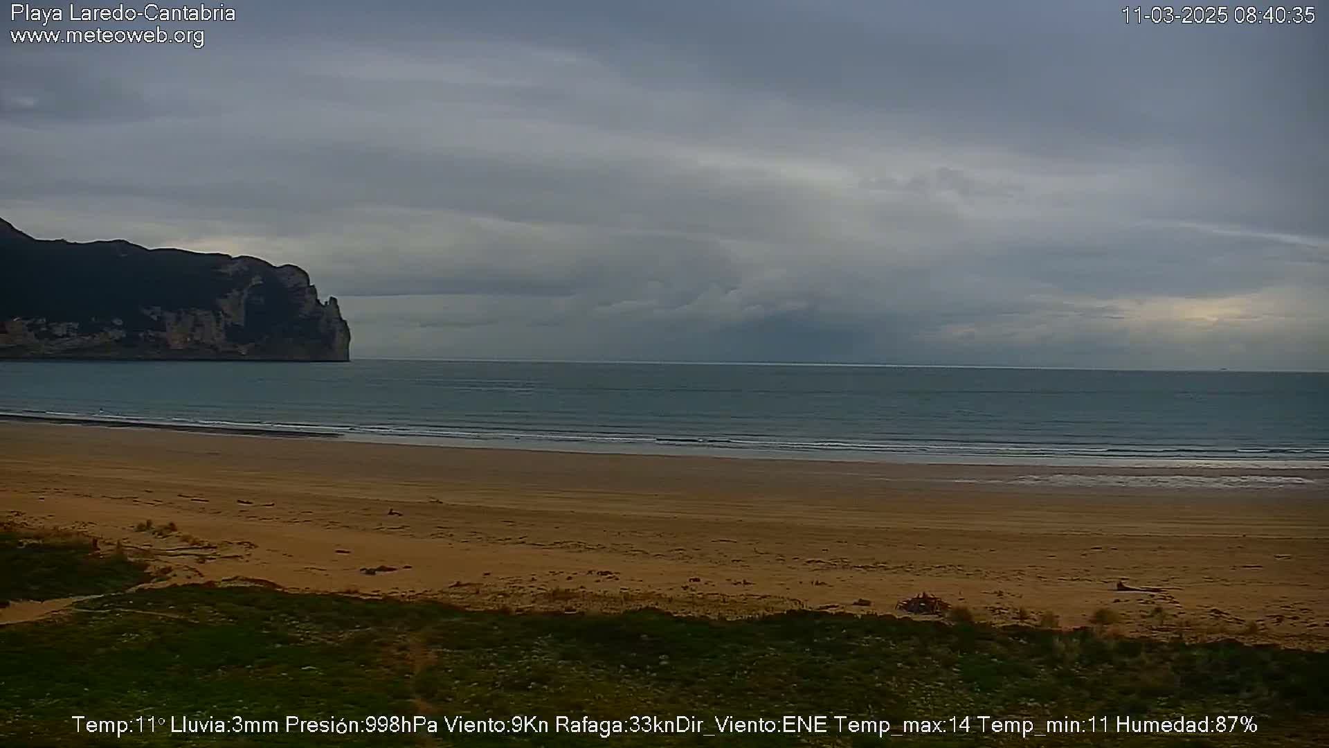 A sandy beach stretches to a calm sea under an overcast sky, with a large cliff visible in the distance.