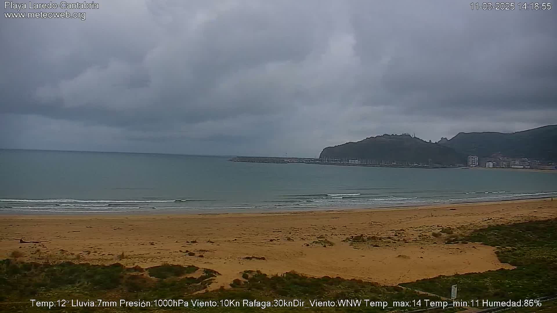 A sandy beach stretches along a calm ocean under an overcast sky, with a distant hill visible in the background.