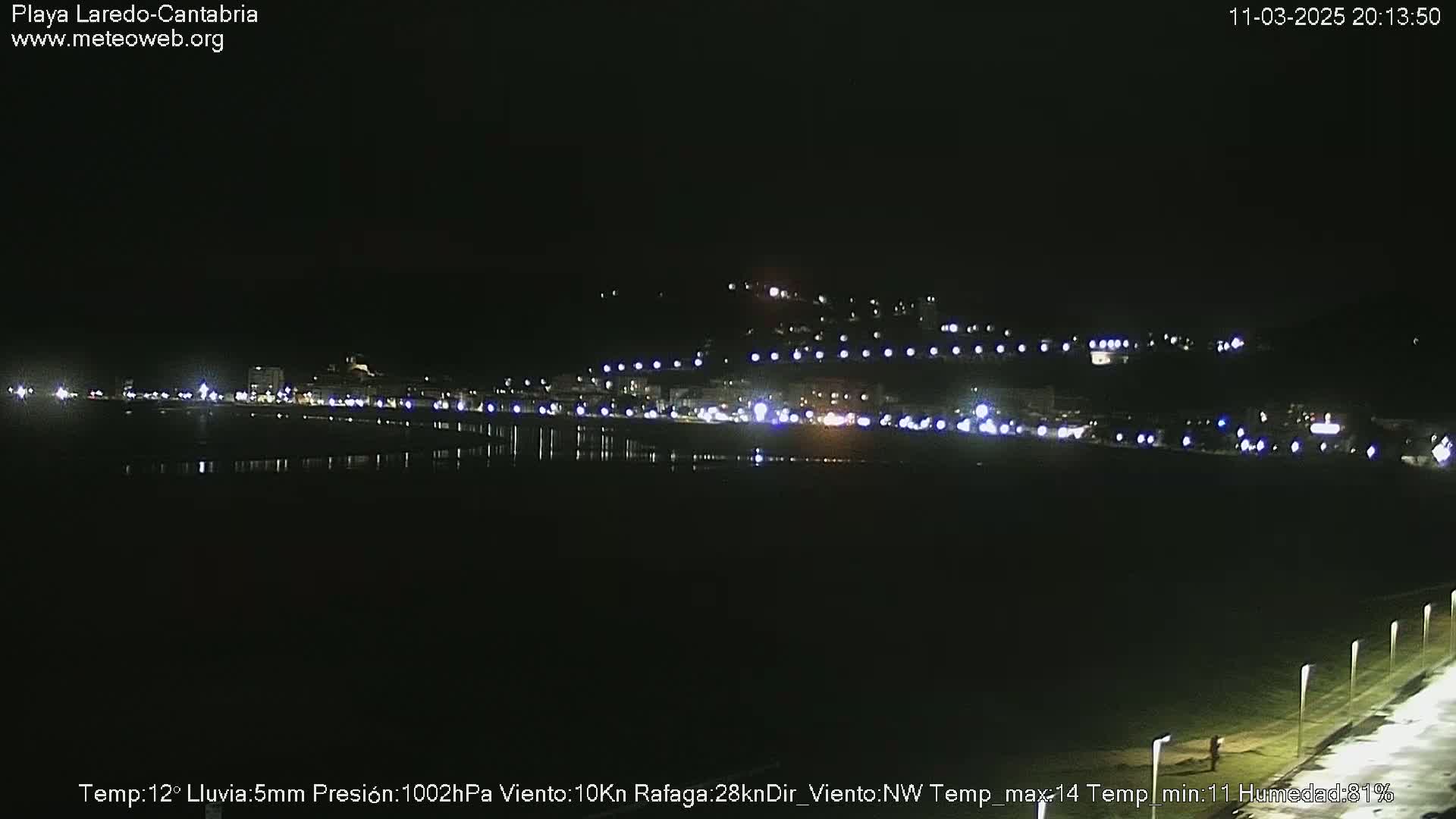 A nighttime coastal scene shows a dark, calm body of water reflecting the lights of a town on a hill in the distance under a clear, dark sky.