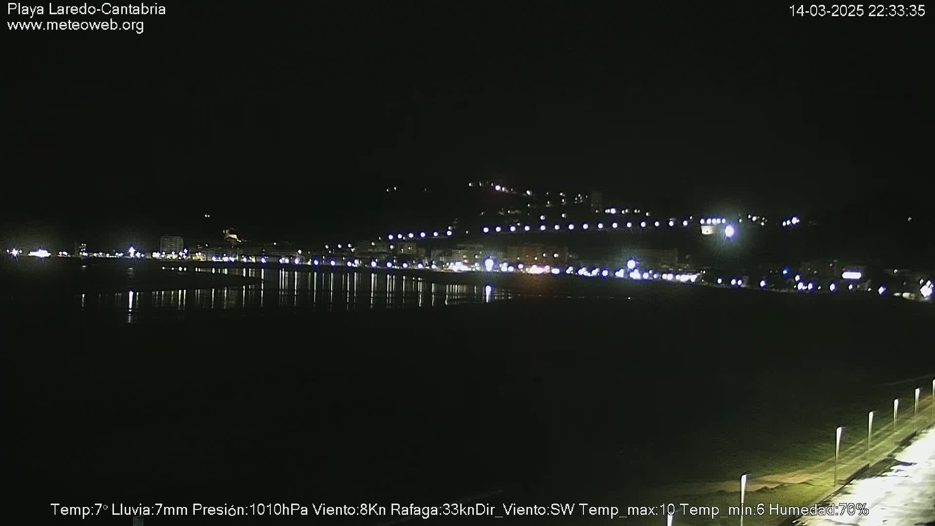 A nighttime view of a coastal town with lights reflecting on calm water under a dark sky.