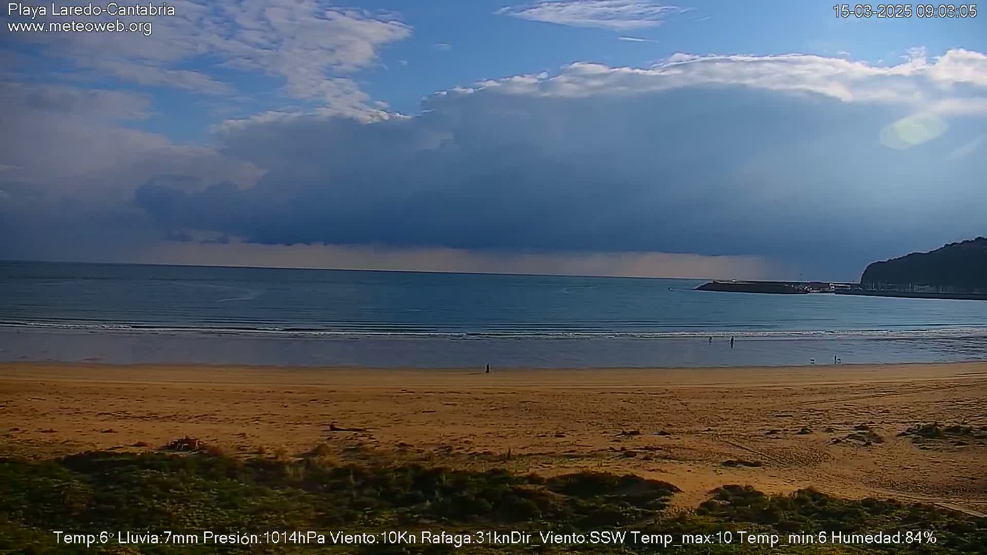 A wide sandy beach stretches toward a calm ocean under a mostly cloudy sky with a dark, ominous cloud formation looming over the horizon.
