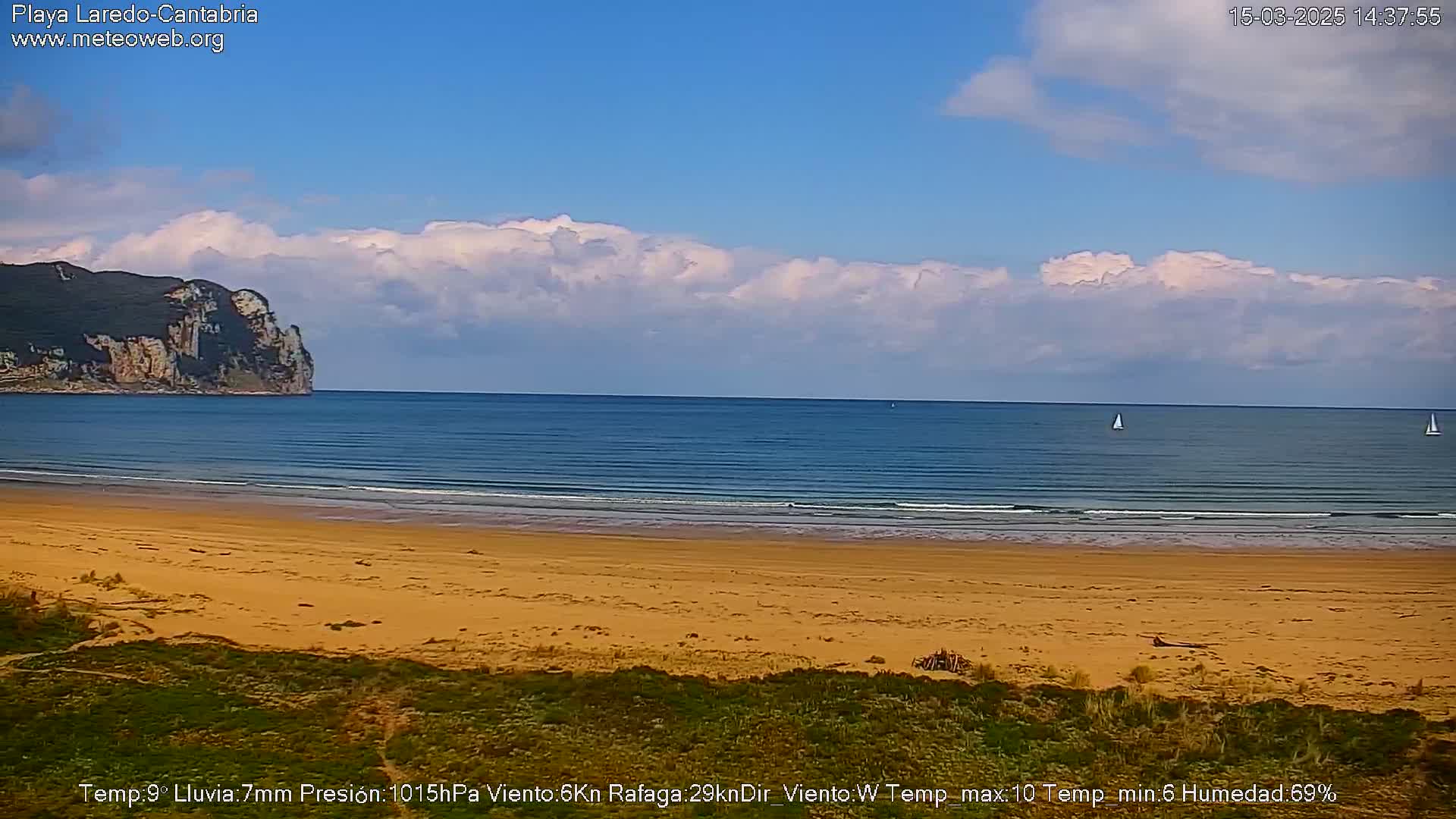 A sandy beach stretches to a calm blue ocean under a partly cloudy sky, with a distant cliff and a couple of sailboats visible on the water.